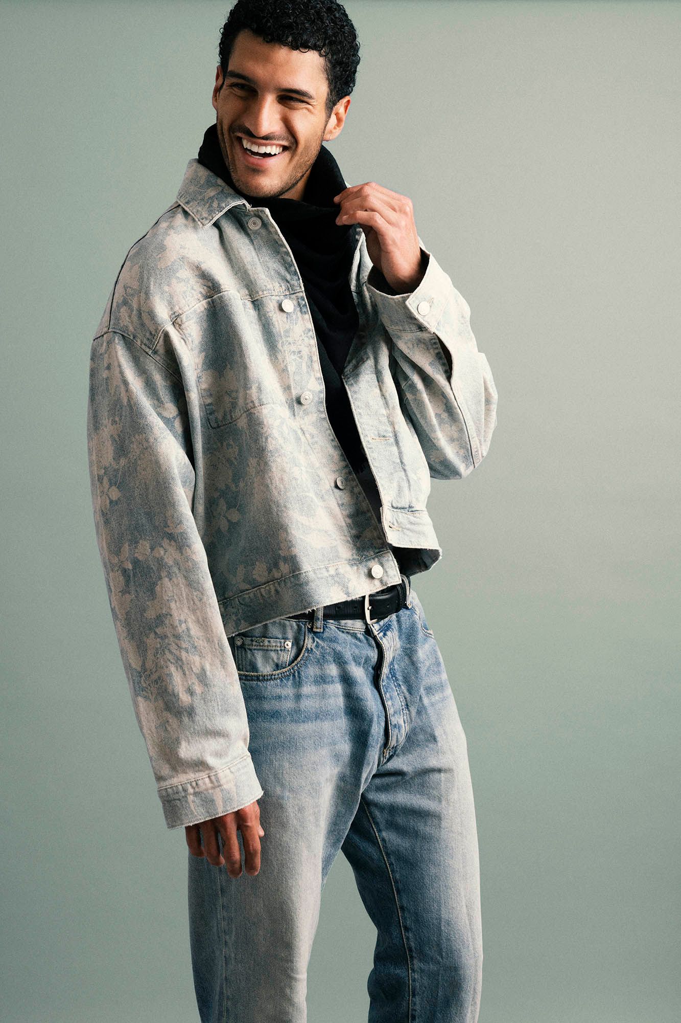 Smiling man in distressed denim jacket and blue jeans posing against gray background.