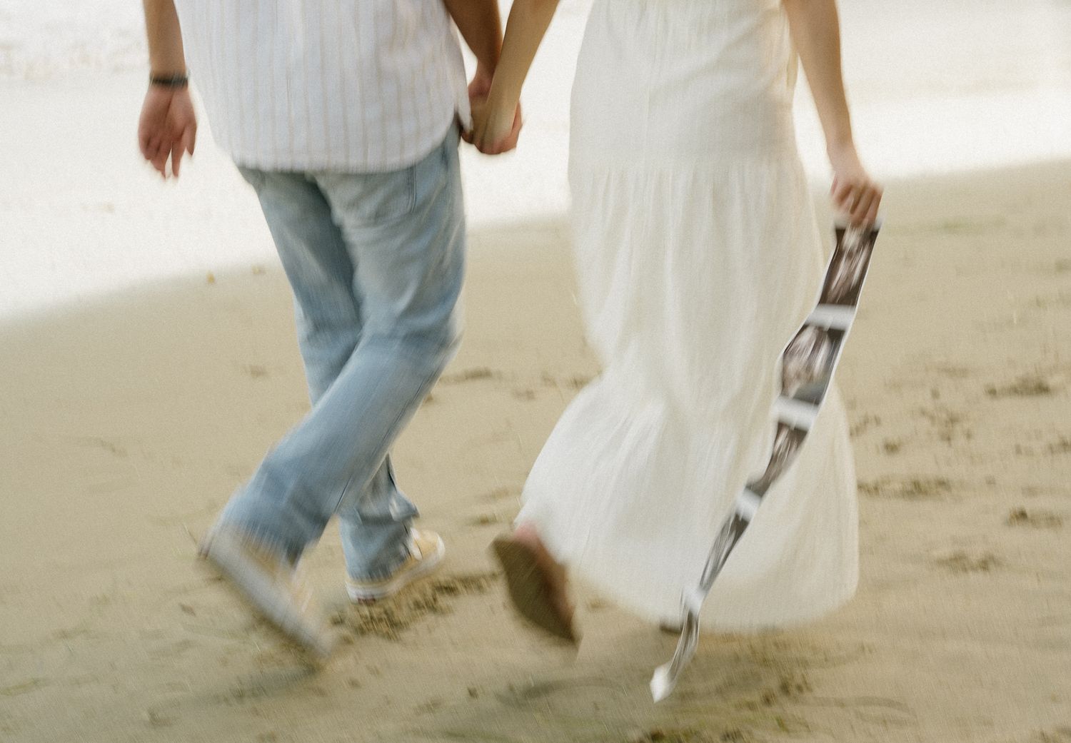 Couple holding hands walking on sandy beach, woman in white dress carrying ultrasound photos.