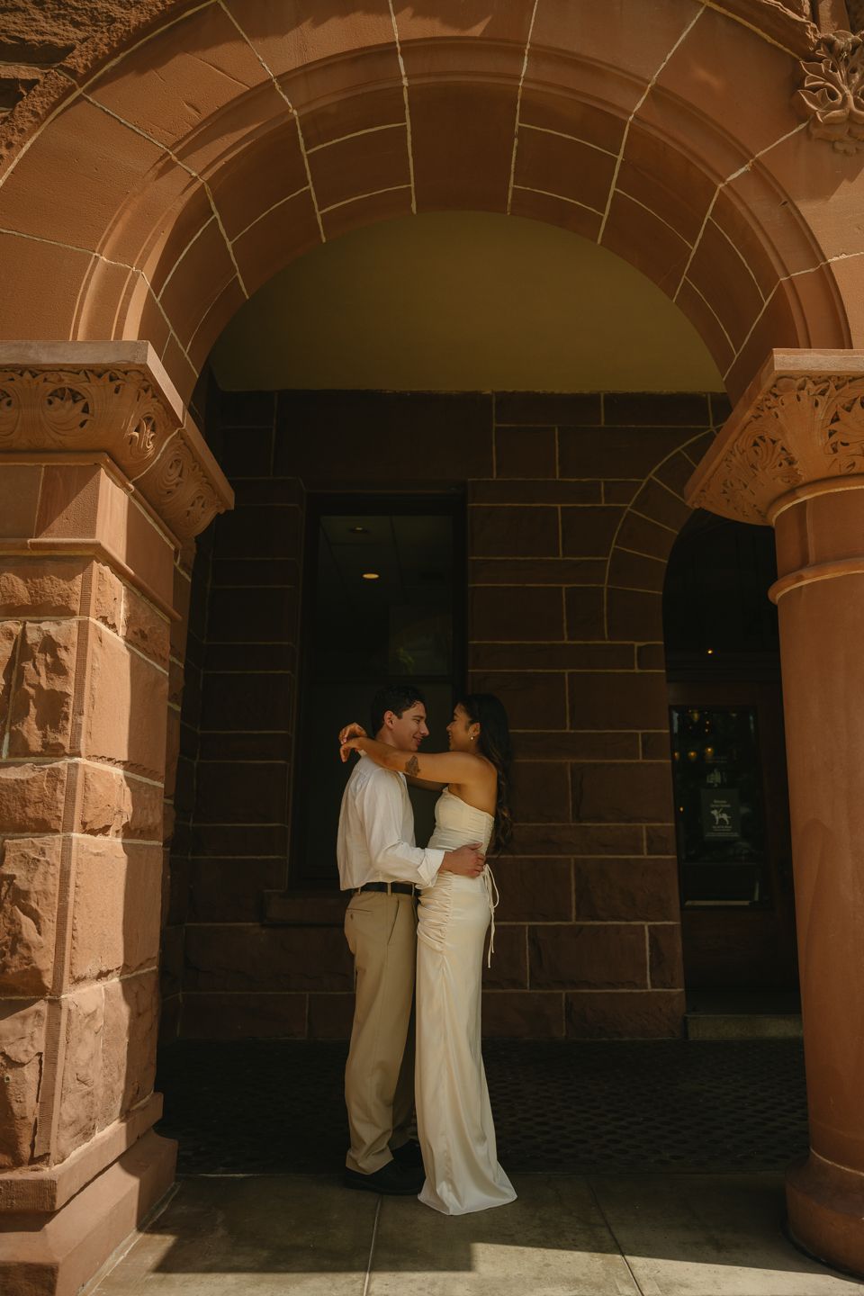 Couple embracing under a stone archway in wedding attire, bride in white gown, groom in tan pants at historic building.