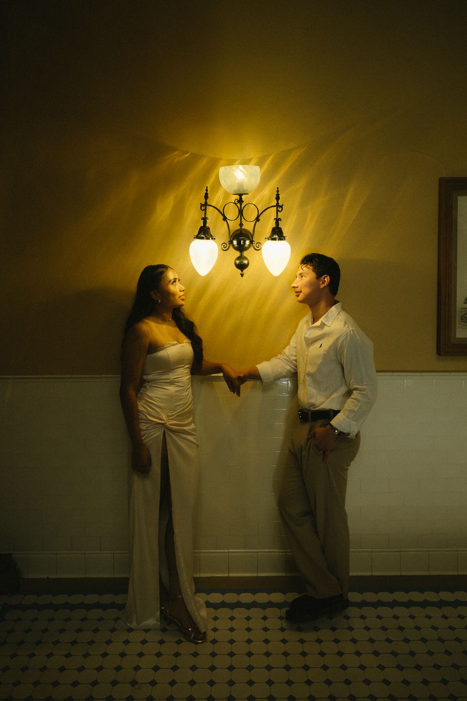 Couple holding hands under warm wall sconce light in elegant interior setting.