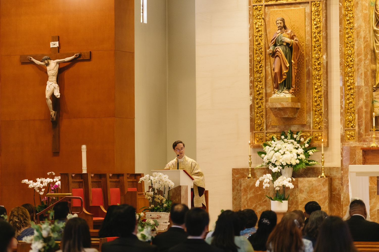 Priest delivering a sermon at a Catholic church altar with a crucifix and congregation seated in pews.