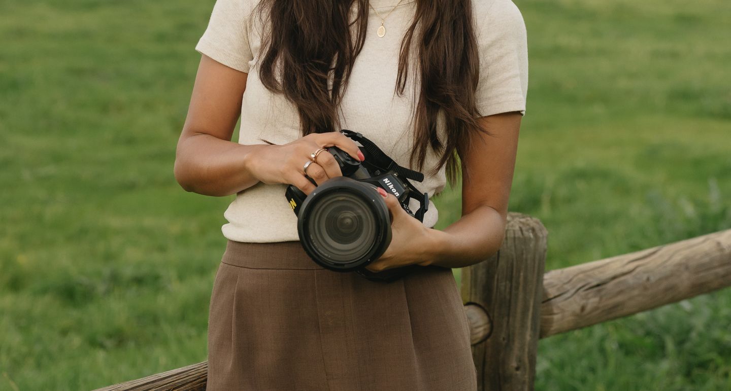 Young woman in cream top and brown skirt holding a camera outdoors by a wooden fence.