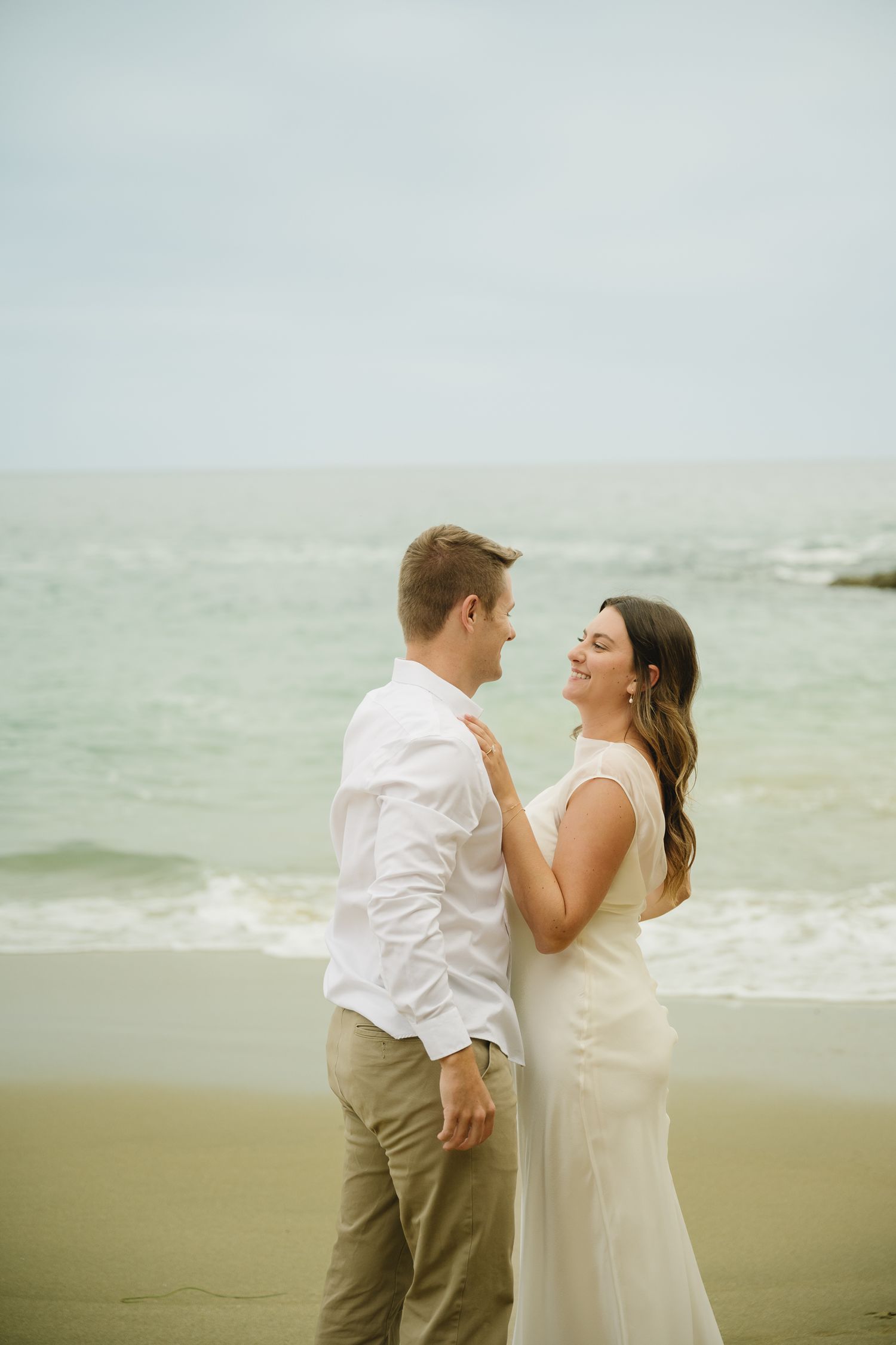 Couple in white attire embracing on a sandy beach with ocean waves in the background.