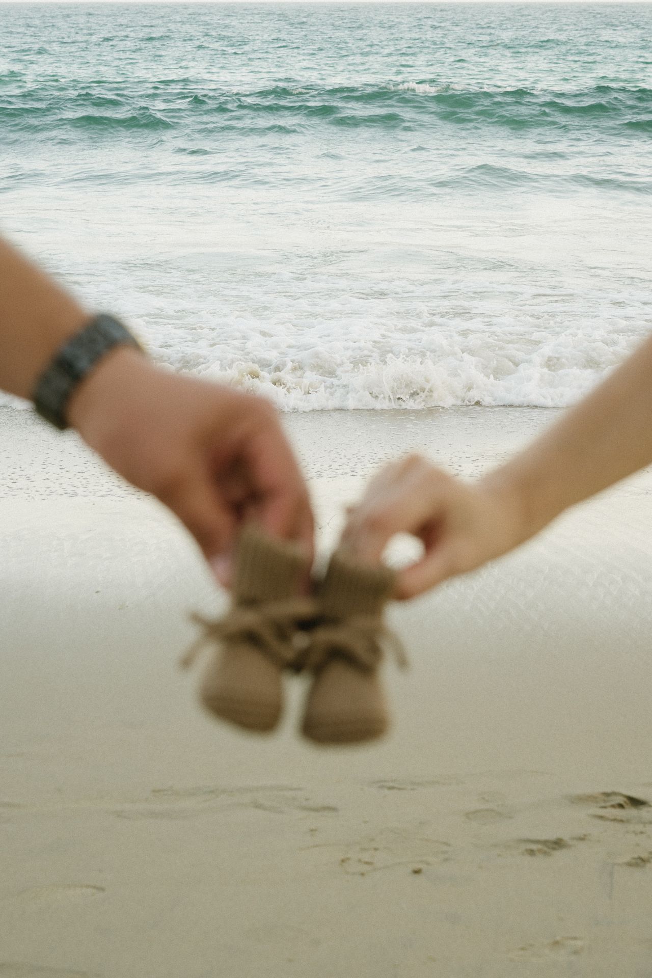 Couple holding tiny baby shoes together on a sandy beach with ocean waves in the background.
