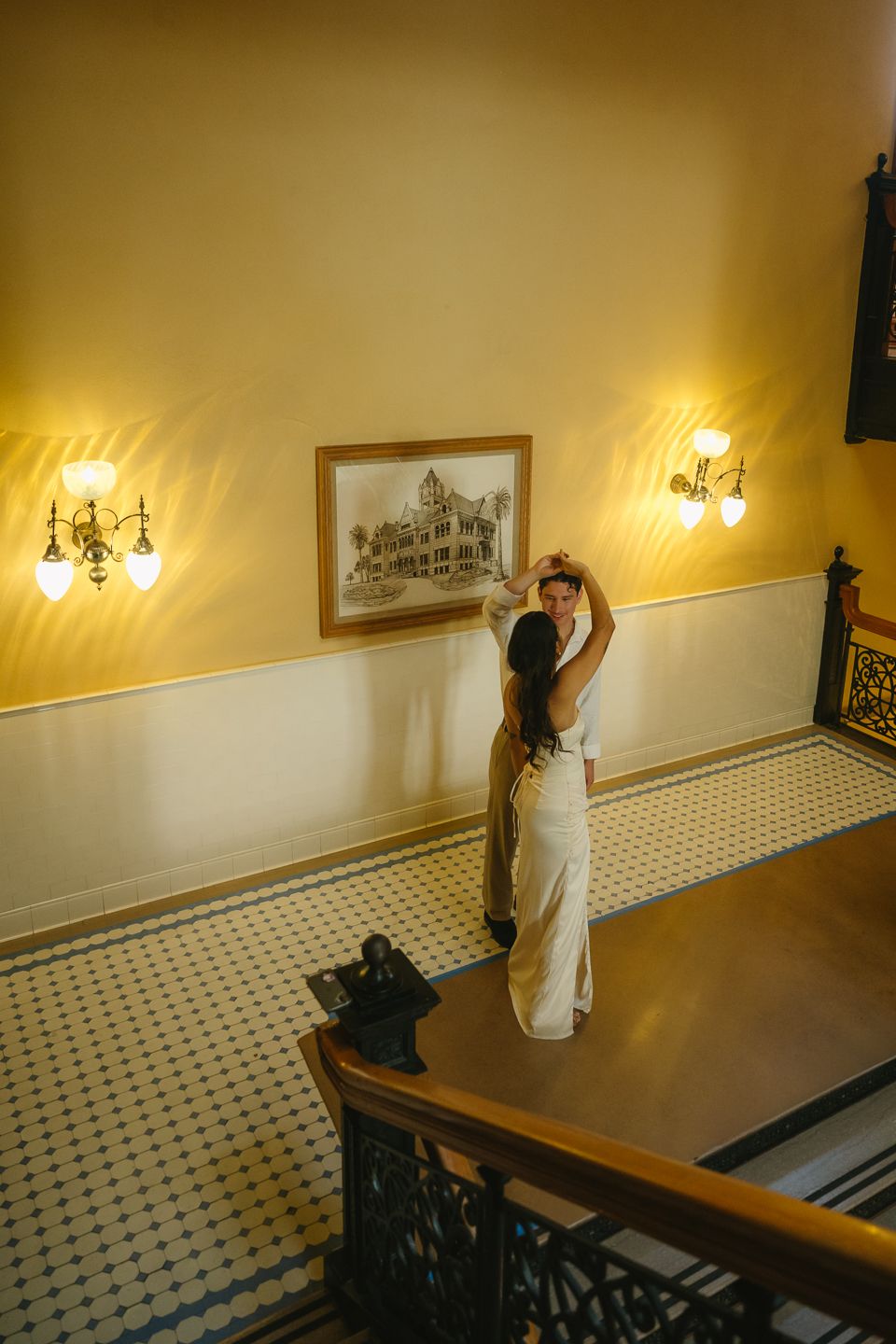 Bride and groom dancing near framed artwork on yellow wall with vintage sconces in elegant historic venue.