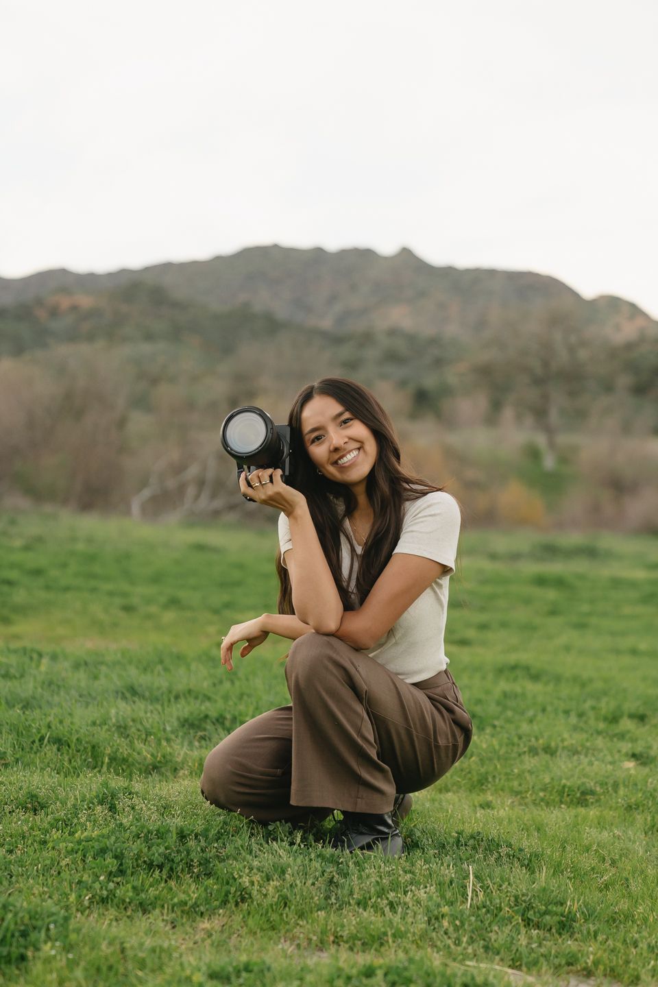 Smiling woman photographer crouching in green field holding DSLR camera with mountains in background.