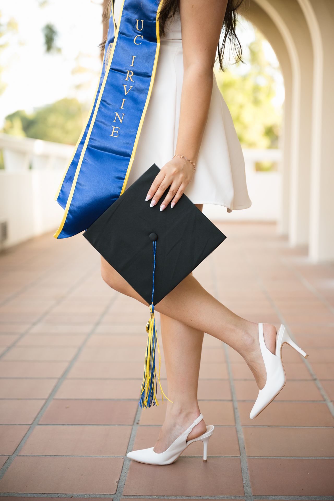 UC Irvine graduate in white dress and heels holding black mortarboard cap with blue and gold tassel.