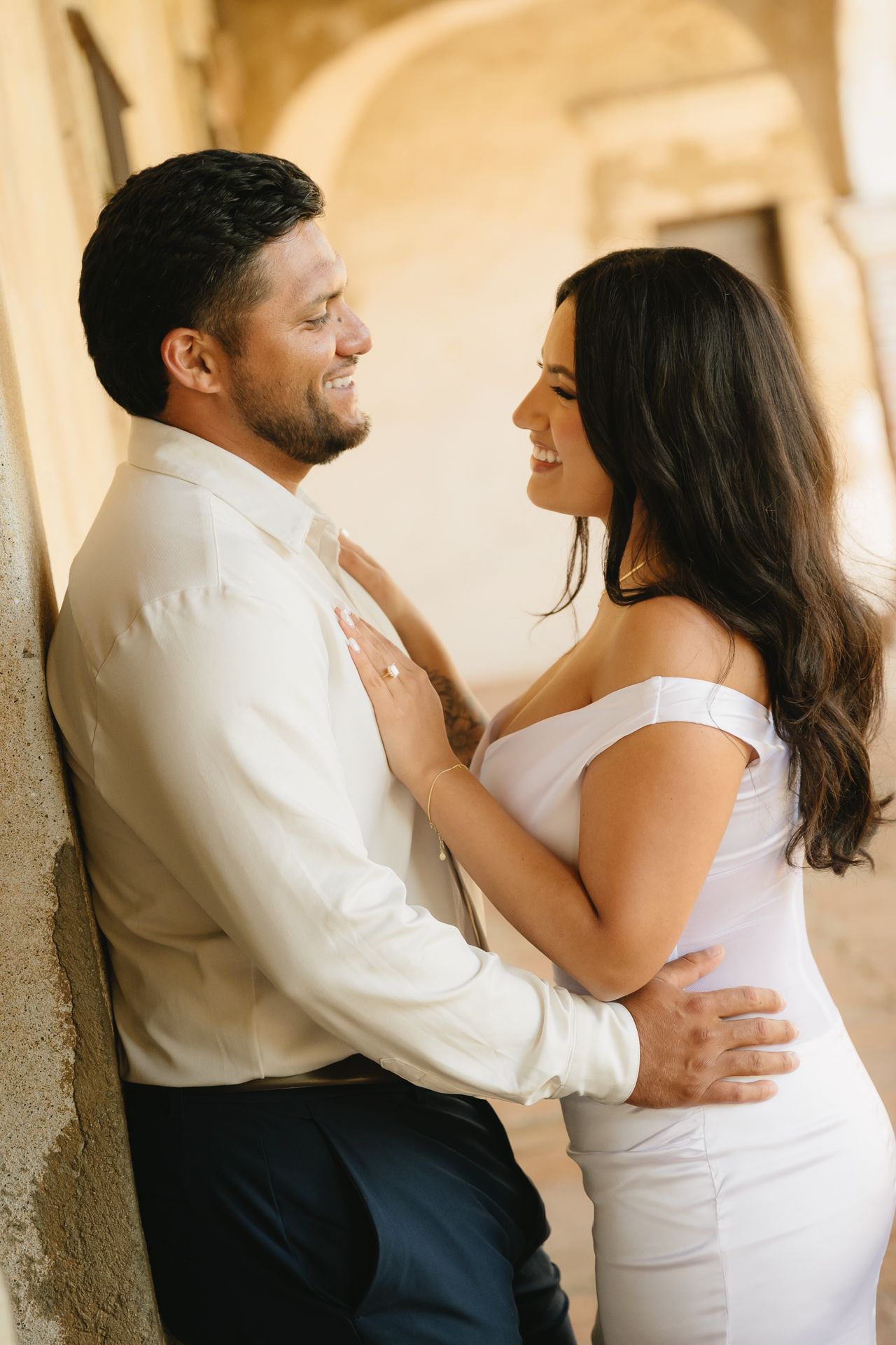 A smiling couple in elegant white attire share a romantic moment leaning against a warm stone wall.