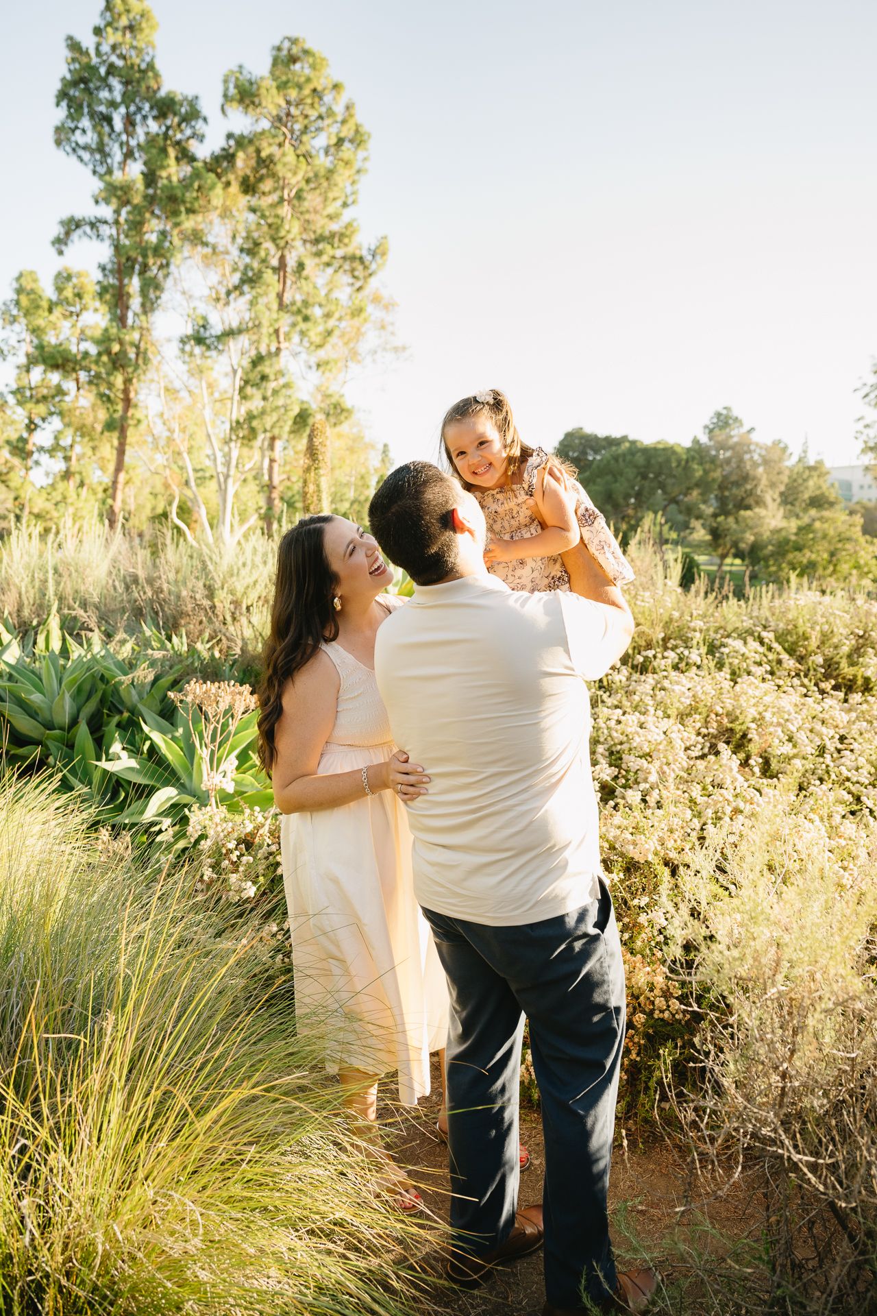 A joyful family of three in a sunlit garden, parents lifting their laughing child among lush greenery.