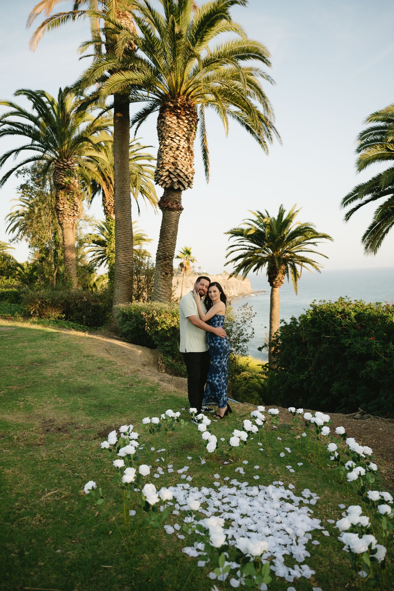 Couple embracing in a garden with white flower petals, palm trees, and ocean view at sunset.