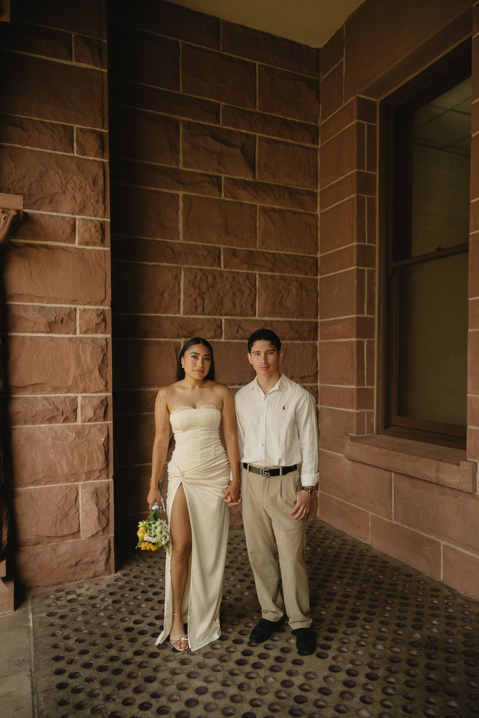 Couple in elegant attire posing before sandstone wall, bride in white slit gown holding bouquet.