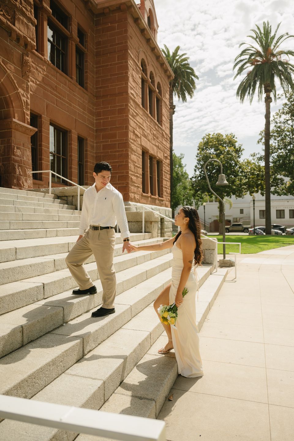 Couple in wedding attire holding hands on steps of a historic red brick building with palm trees.