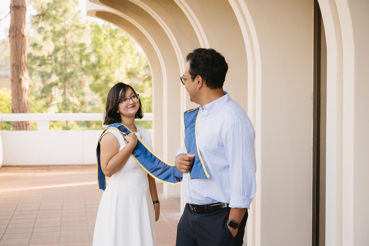 Couple smiling and holding hands under arched colonnade outdoors, woman in white dress, man in blue tie.