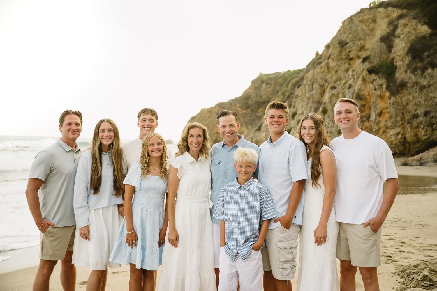 Family of nine posing together on a sandy beach with rocky cliffs in the background on a sunny day.
