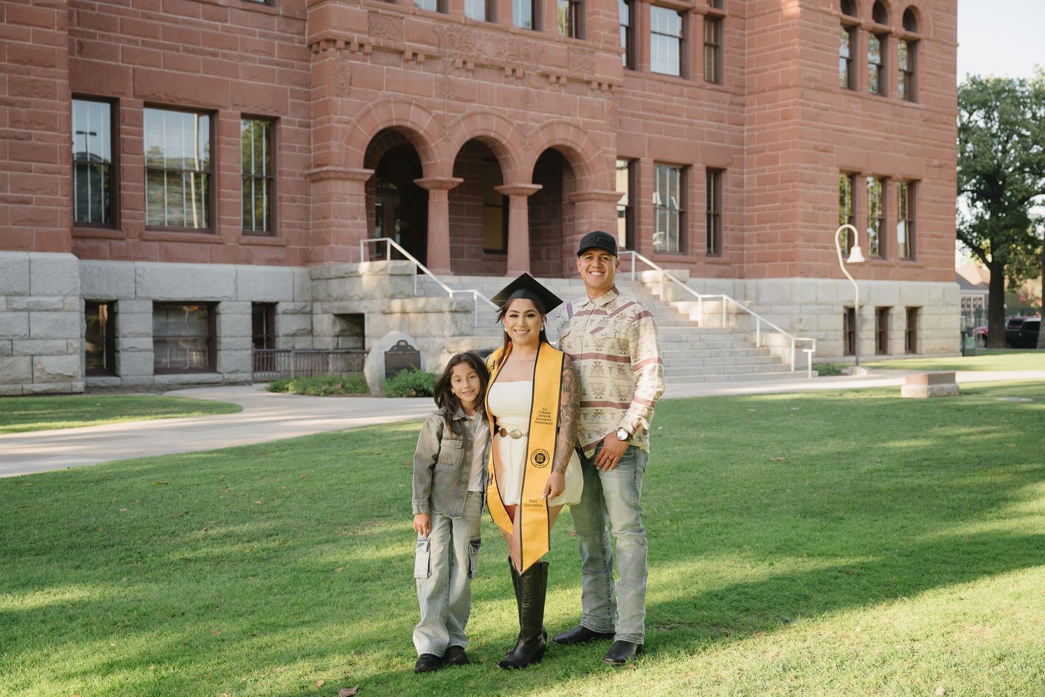 Graduate in cap and gown poses with family in front of brick university building on green lawn.