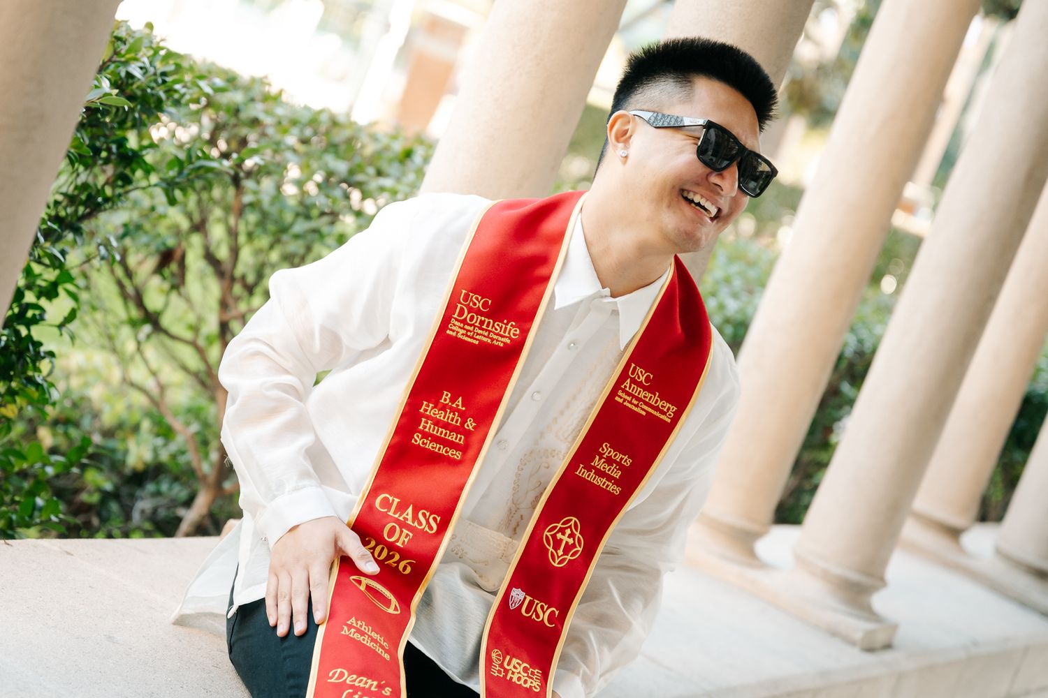 USC graduate smiling in red graduation sash and sunglasses, posing outdoors near white columns.