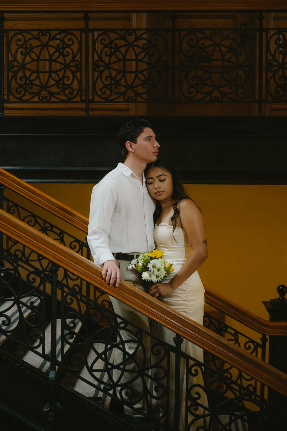 Couple in wedding attire posing on ornate wooden staircase, bride holding yellow and white bouquet.
