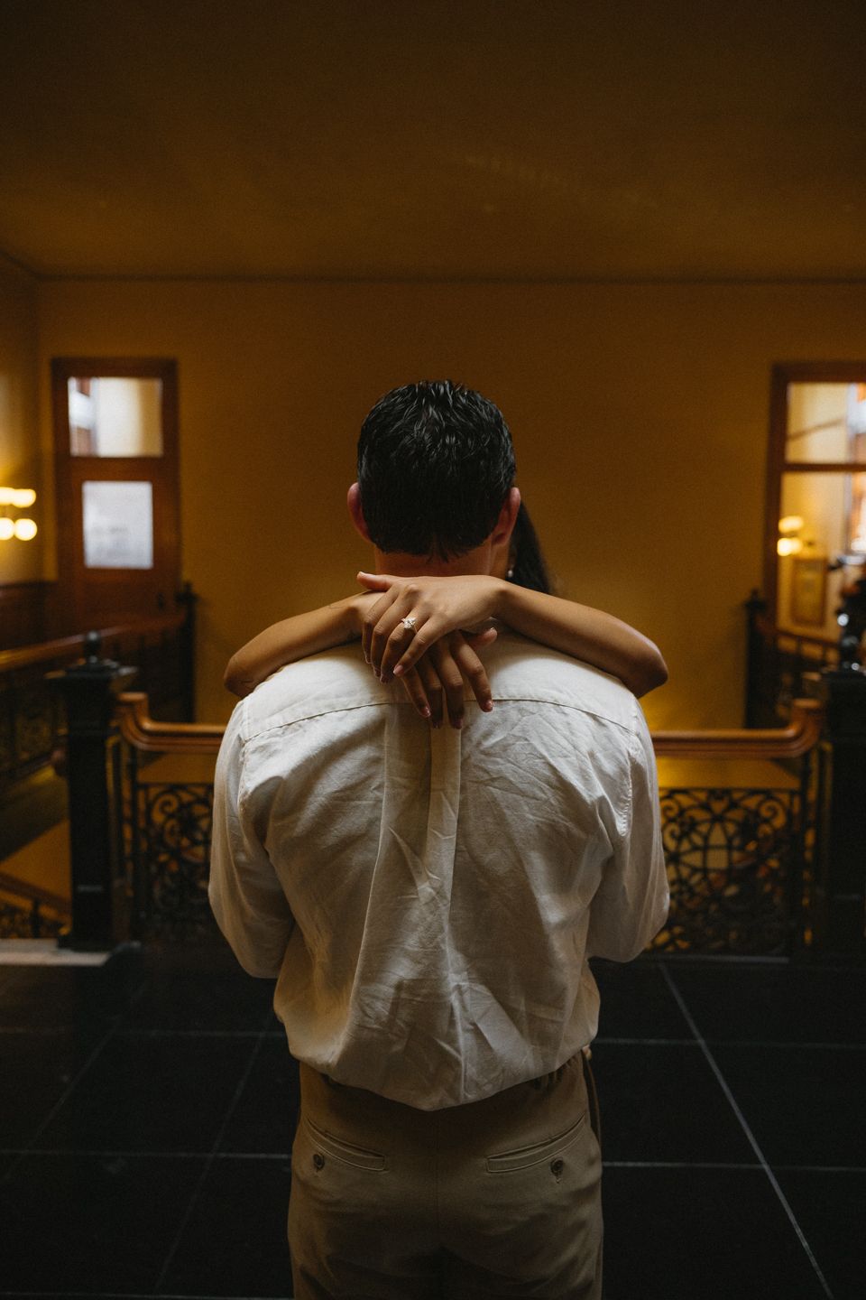 A person in a white shirt being embraced from behind by another person in a warmly lit interior.