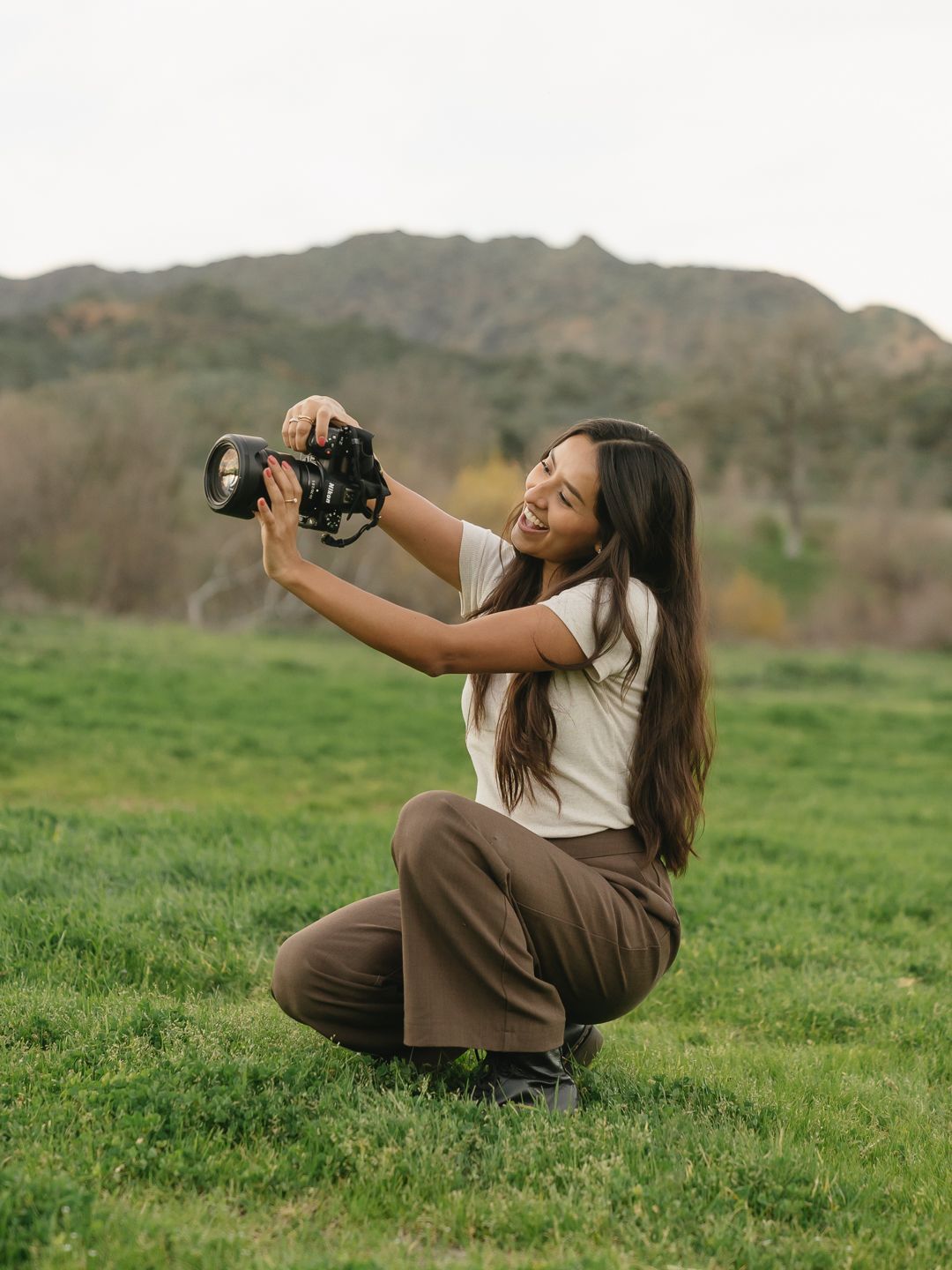 Woman crouching on green grass holding a camera up, with rolling hills and mountains in the background.
