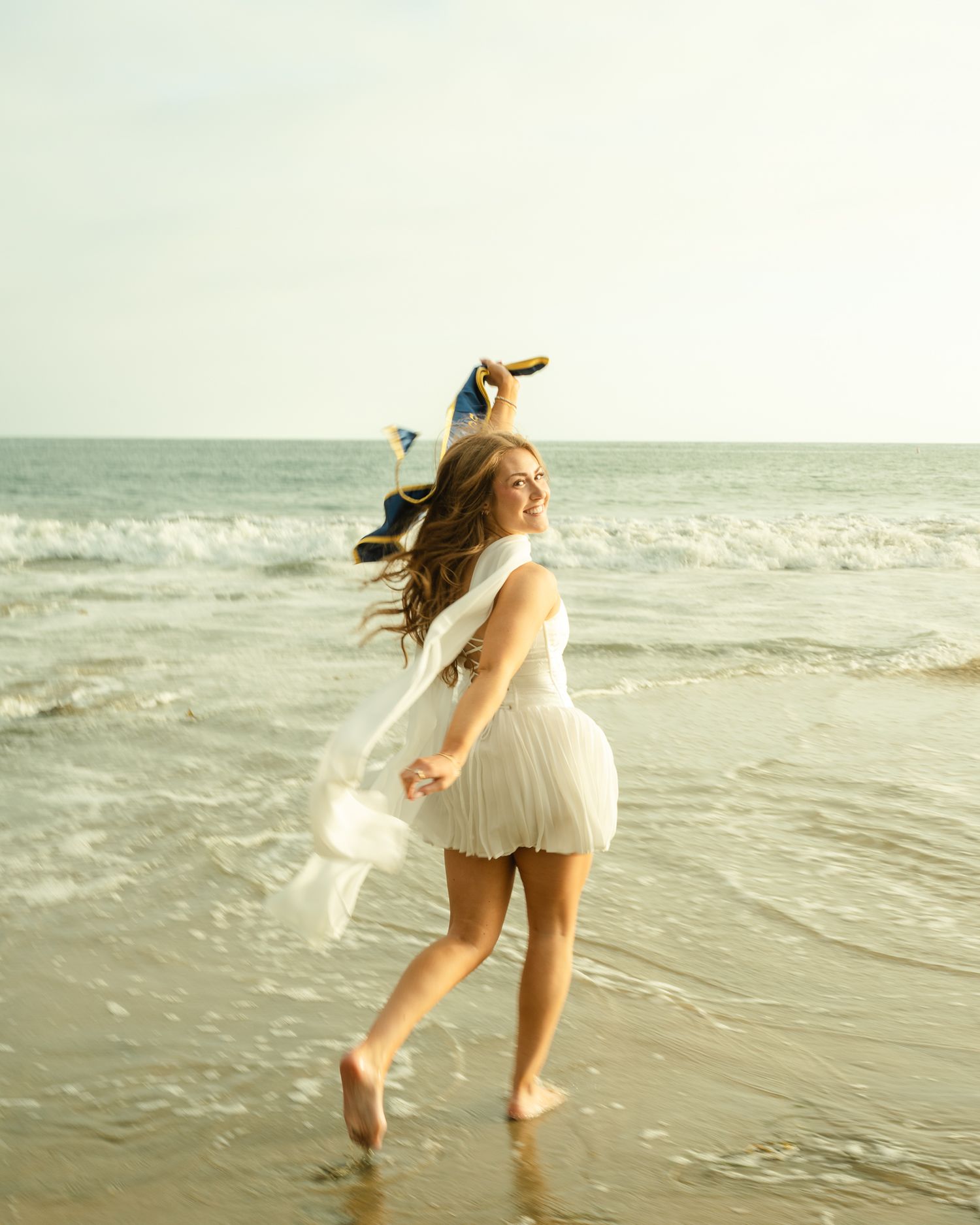 A joyful woman in a white dress twirls barefoot on a sunny beach, her long hair flowing in the ocean breeze.