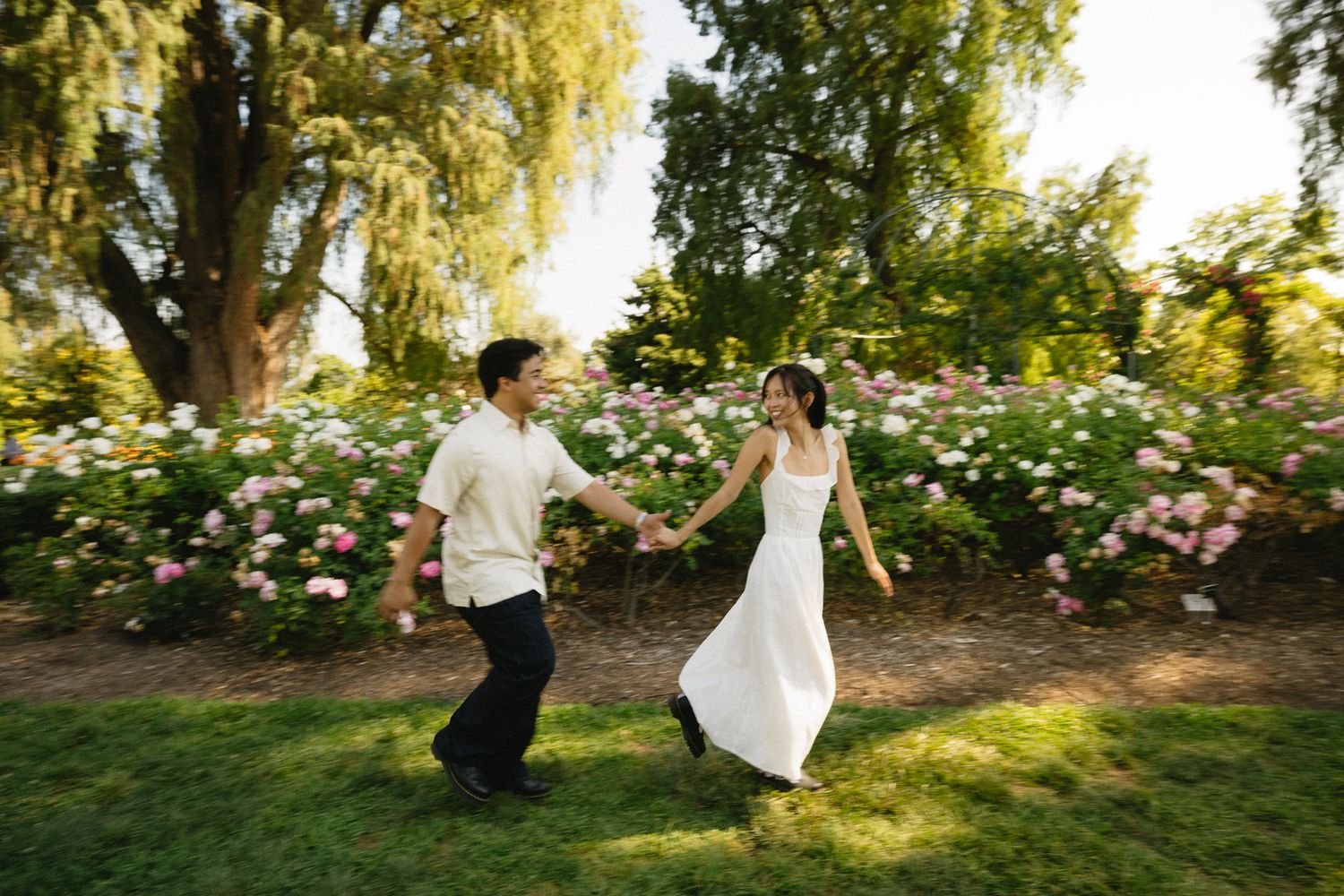 A joyful couple holds hands and dances in a lush garden with pink roses, the bride in a white gown.