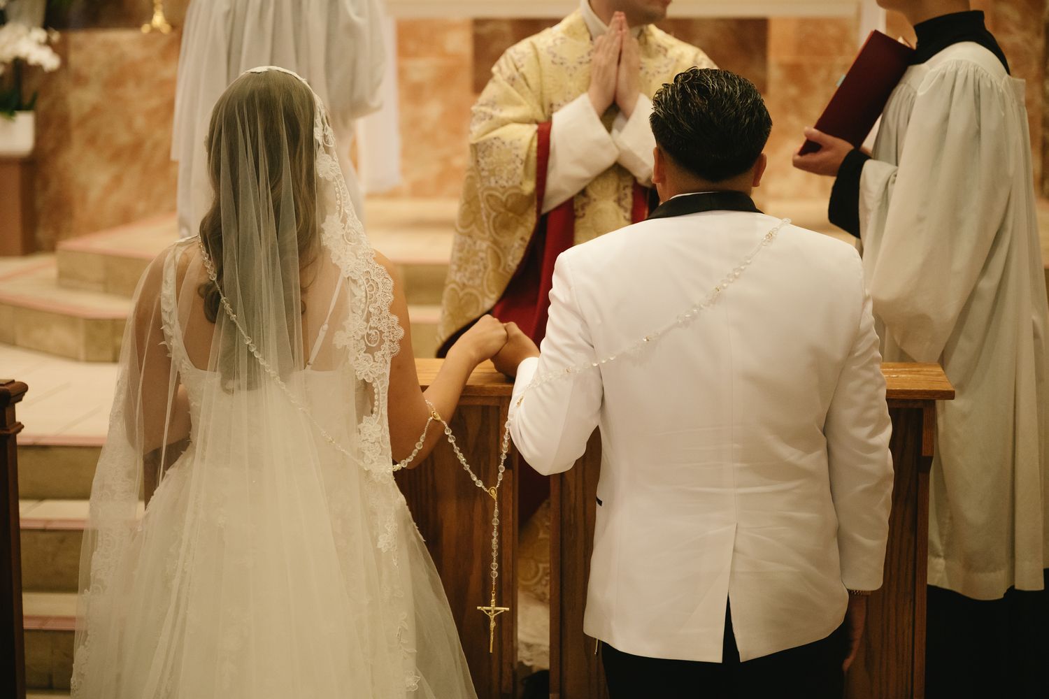 Bride and groom kneeling at church altar during Catholic wedding ceremony with priest praying behind them.