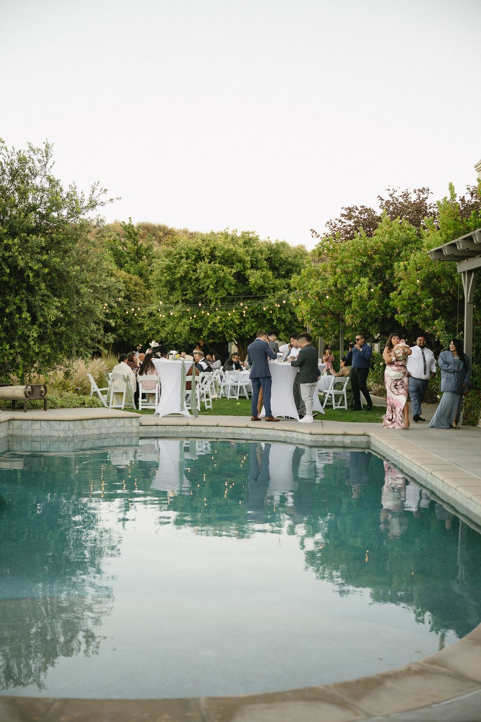 Elegant outdoor wedding reception beside a pool with string lights, and guests mingling among lush greenery located in Inland Empire.