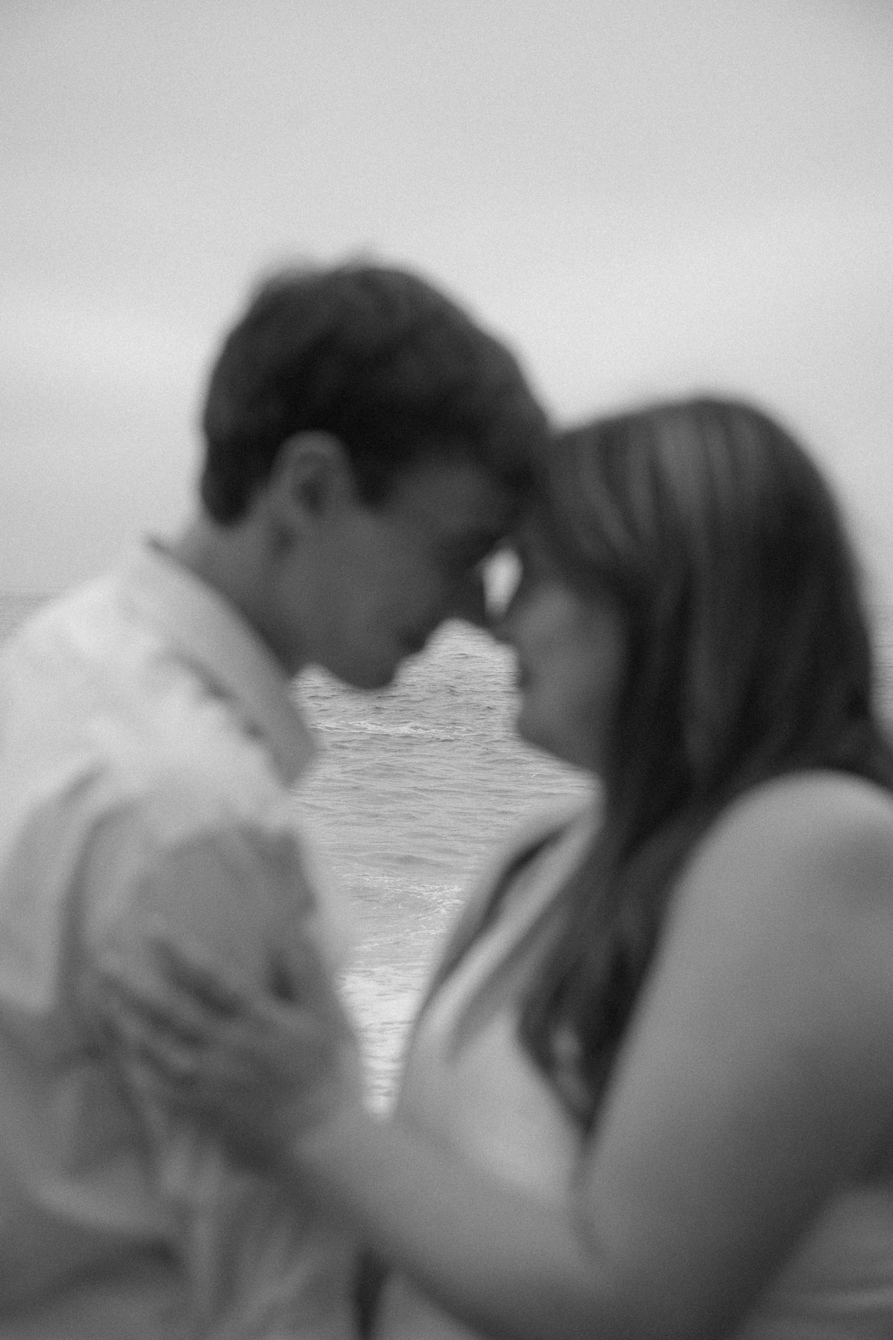 Black and white photo of a romantic couple holding hands and touching foreheads near the ocean.