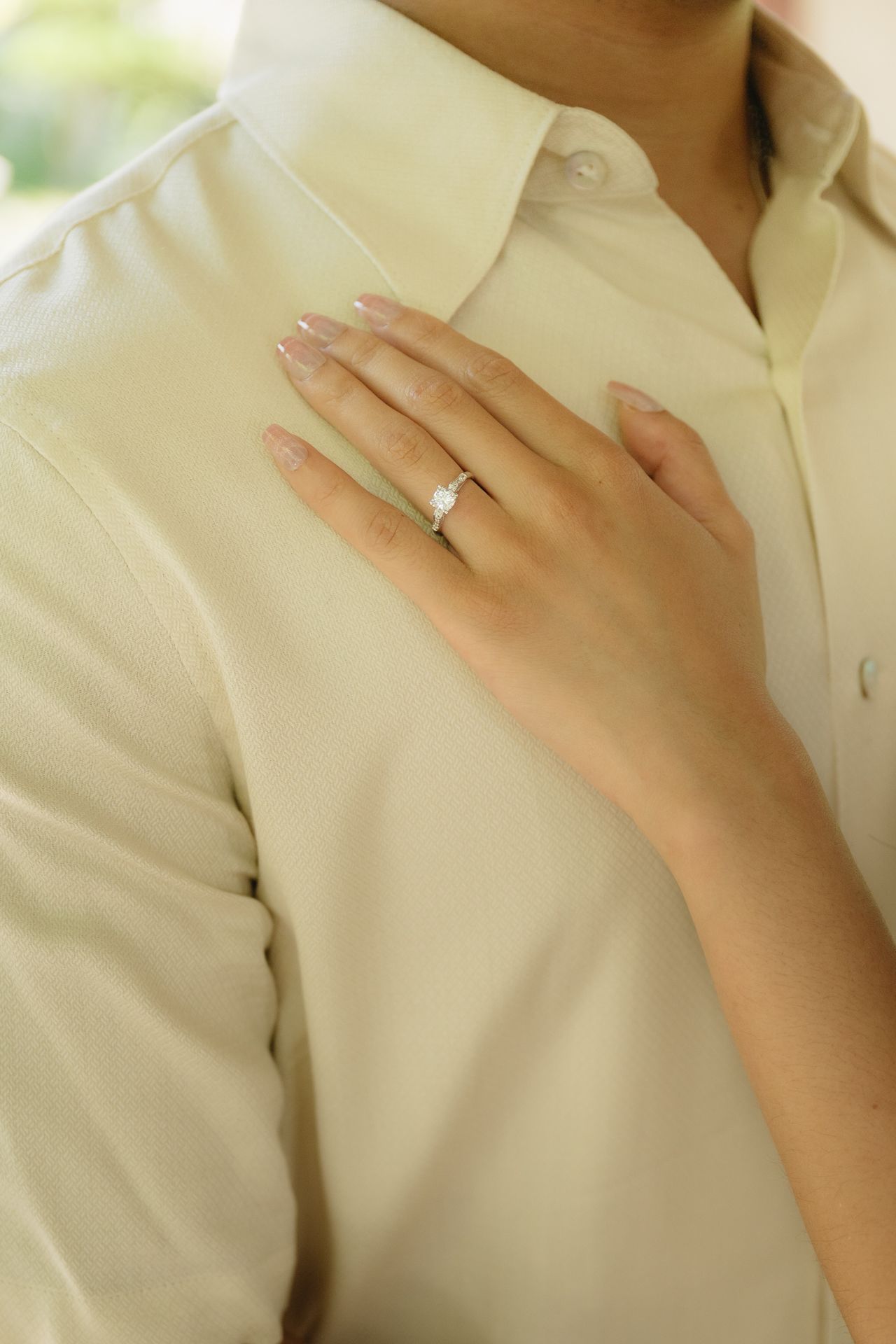 Close-up of a diamond solitaire ring on a woman's hand resting on a cream button-up shirt.