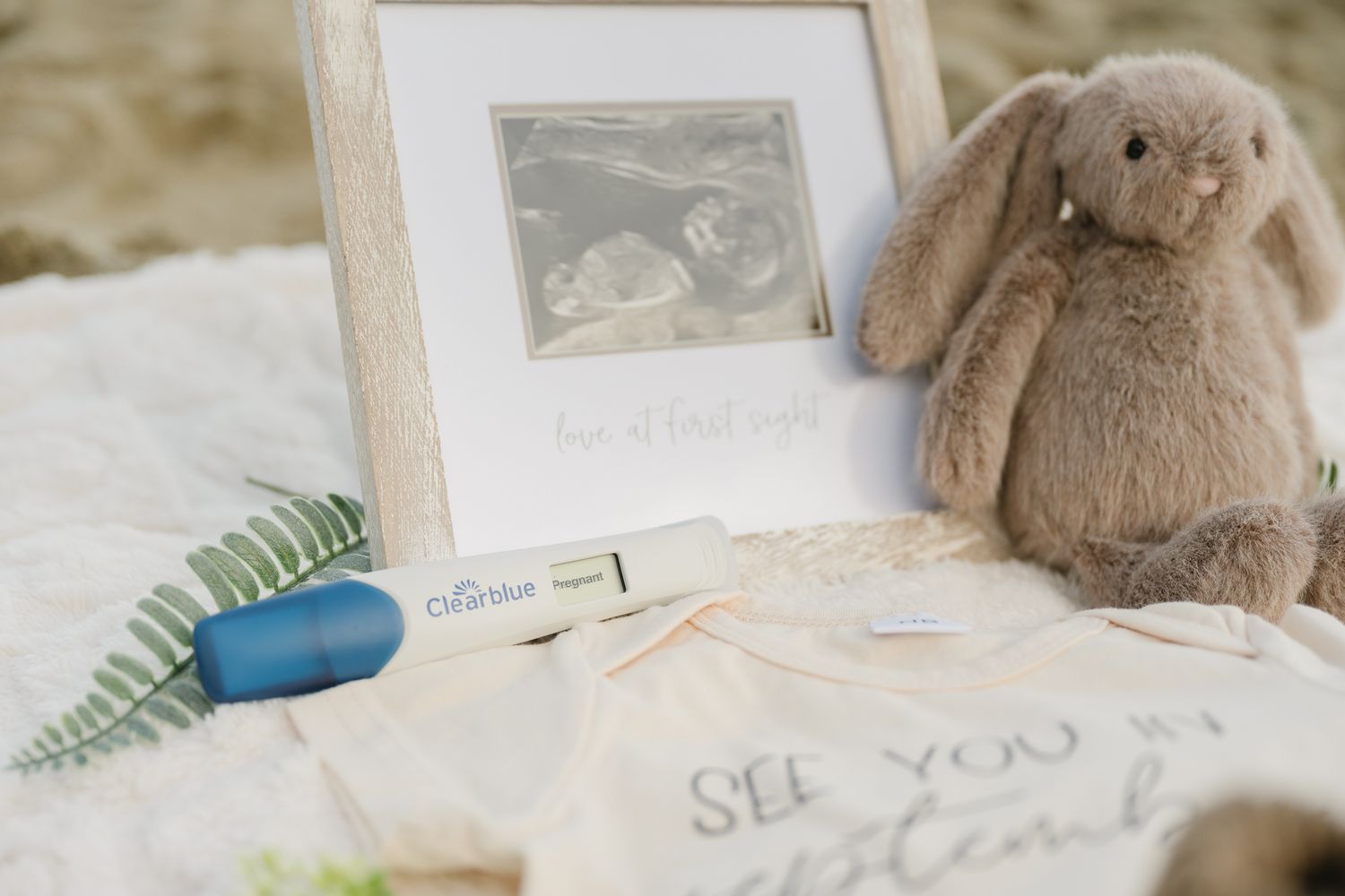 Pregnancy announcement flatlay with ultrasound photo, Clearblue test, stuffed bunny, and baby onesie on white blanket.