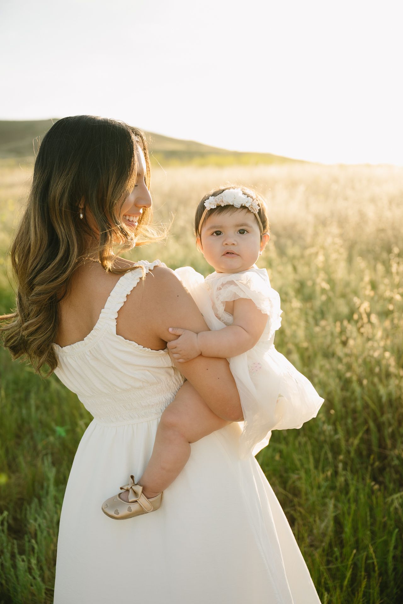 Mother in white dress holding baby girl in white outfit and floral headband in a sunlit green field.