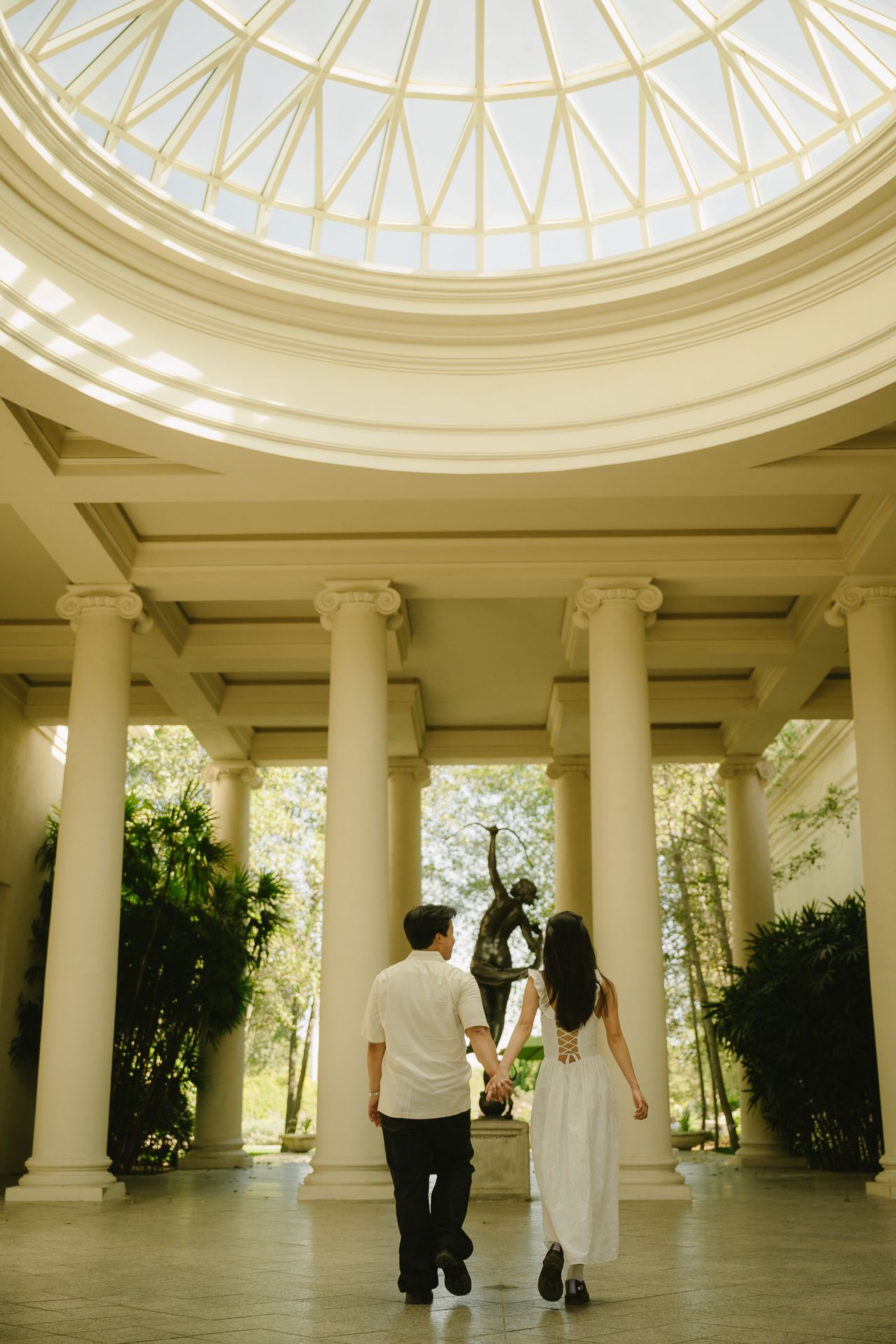 Couple holding hands walking toward classical columns under a domed skylight in an elegant rotunda.