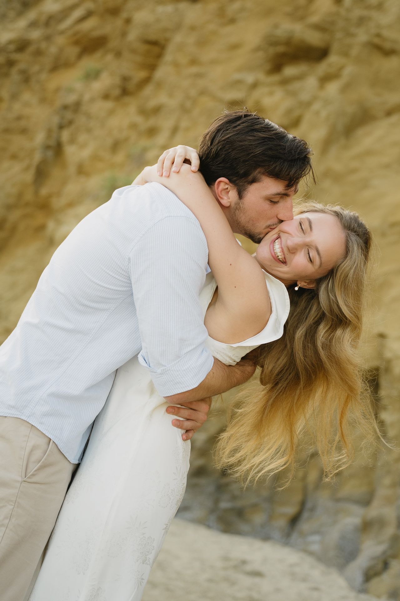 A couple embraces on a sandy beach, man kissing smiling blonde woman in white dress near rocky cliffs.