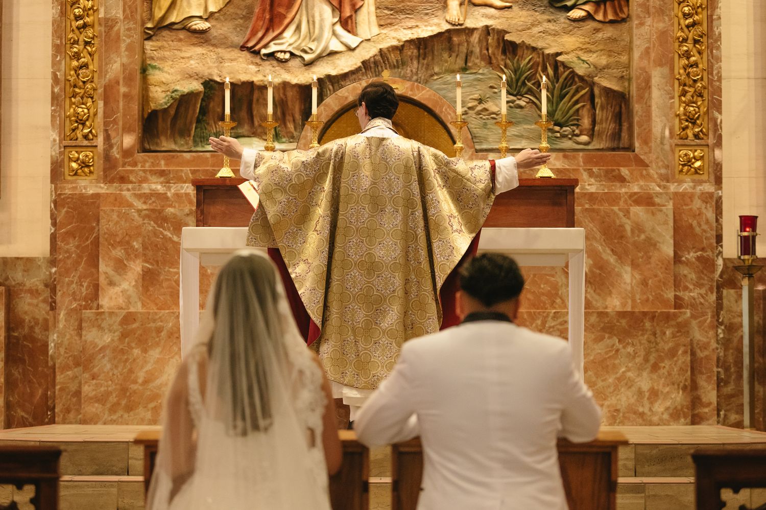 Bride and groom kneeling before a priest at a Catholic church altar during a traditional wedding ceremony.