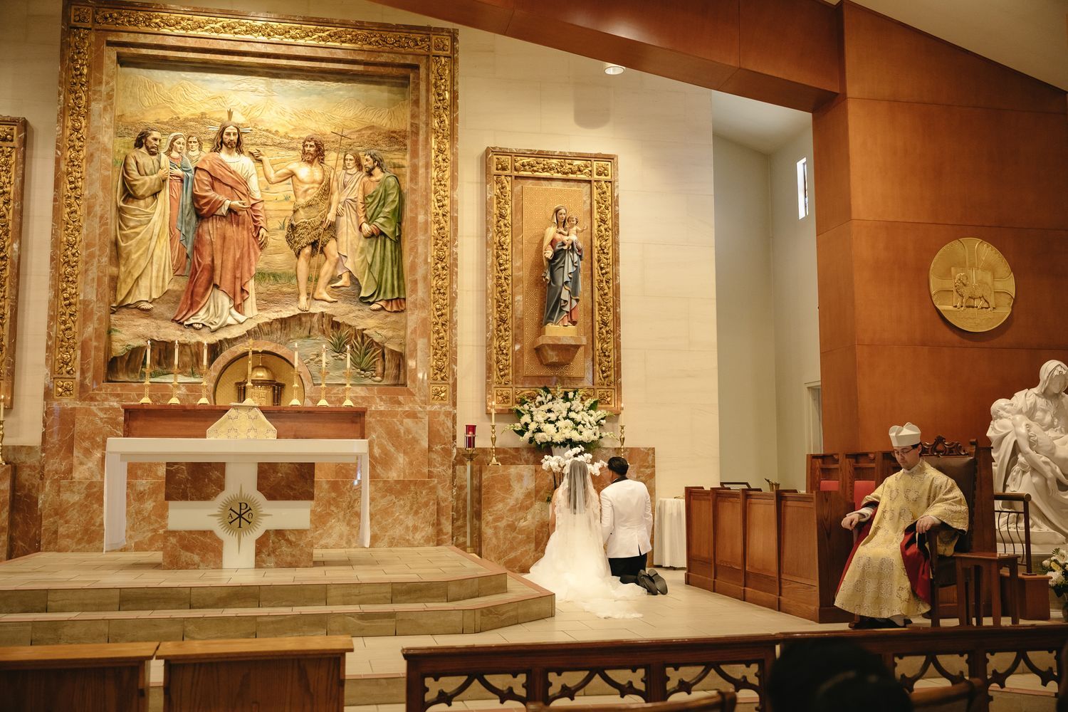 Bride kneeling at church altar during Catholic wedding ceremony with ornate religious artwork and priest officiating.