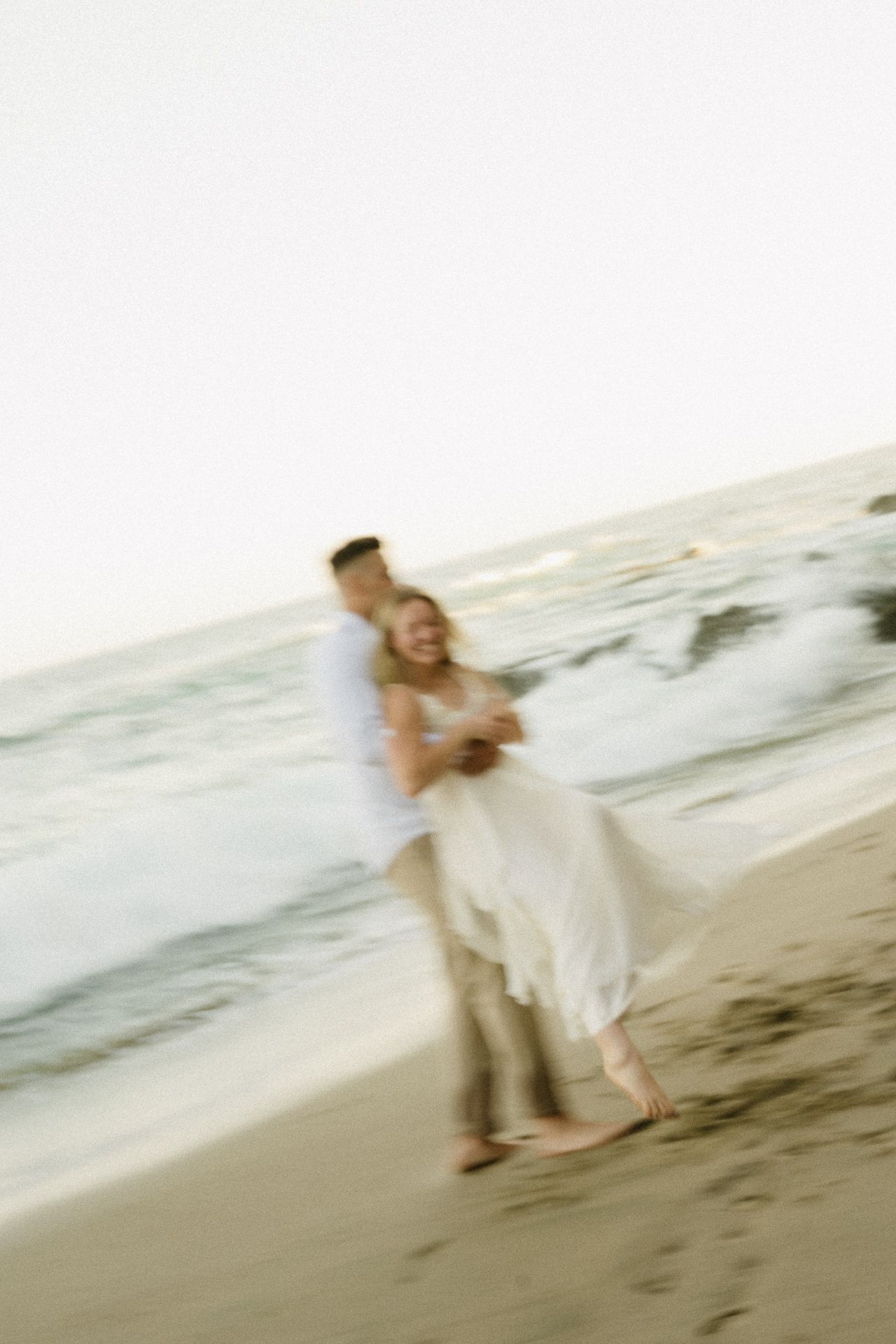 A couple embraces and dances joyfully on a sandy beach near ocean waves in a dreamy, motion-blurred romantic photo.