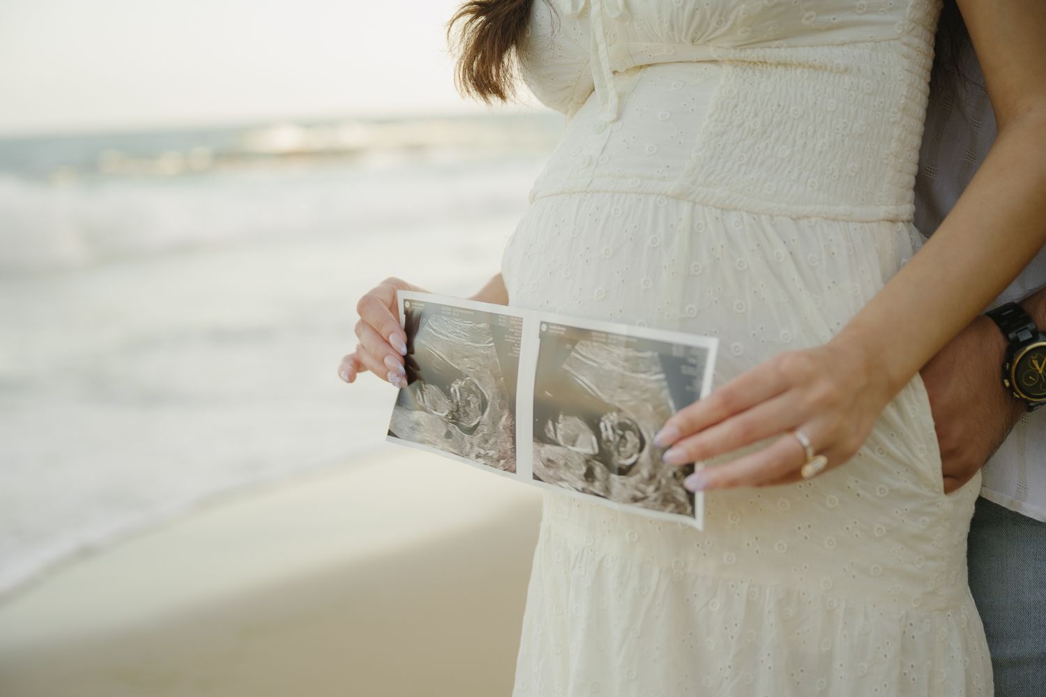 Pregnant woman in white dress holding ultrasound photos on a beach, with ocean waves in the background.