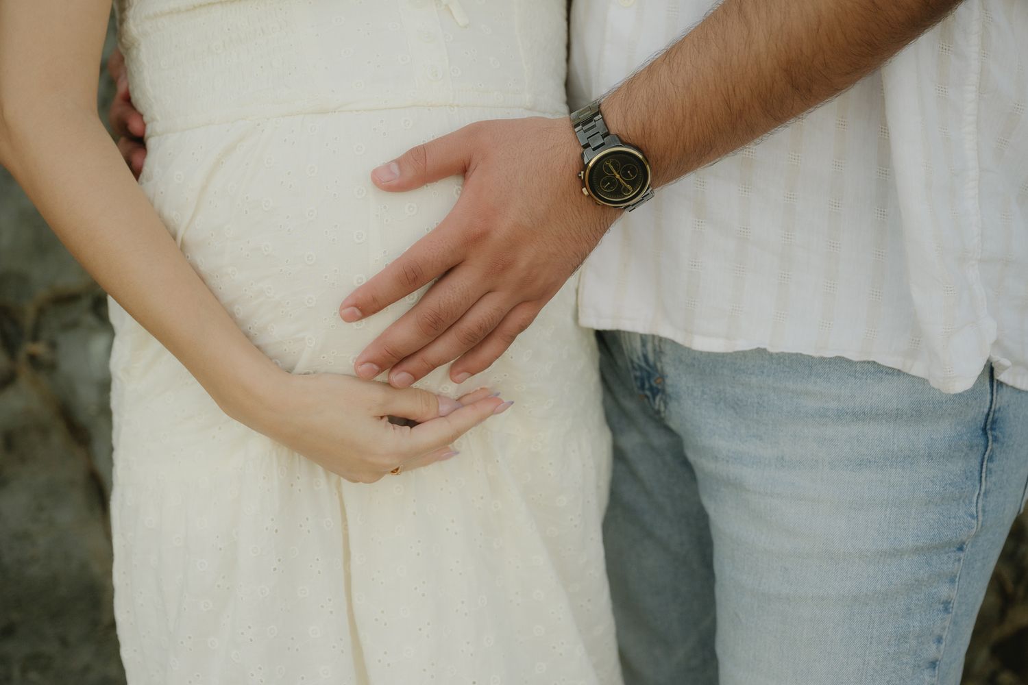 Pregnant woman in white dress with partner's hand on baby bump, maternity couple photo outdoors.