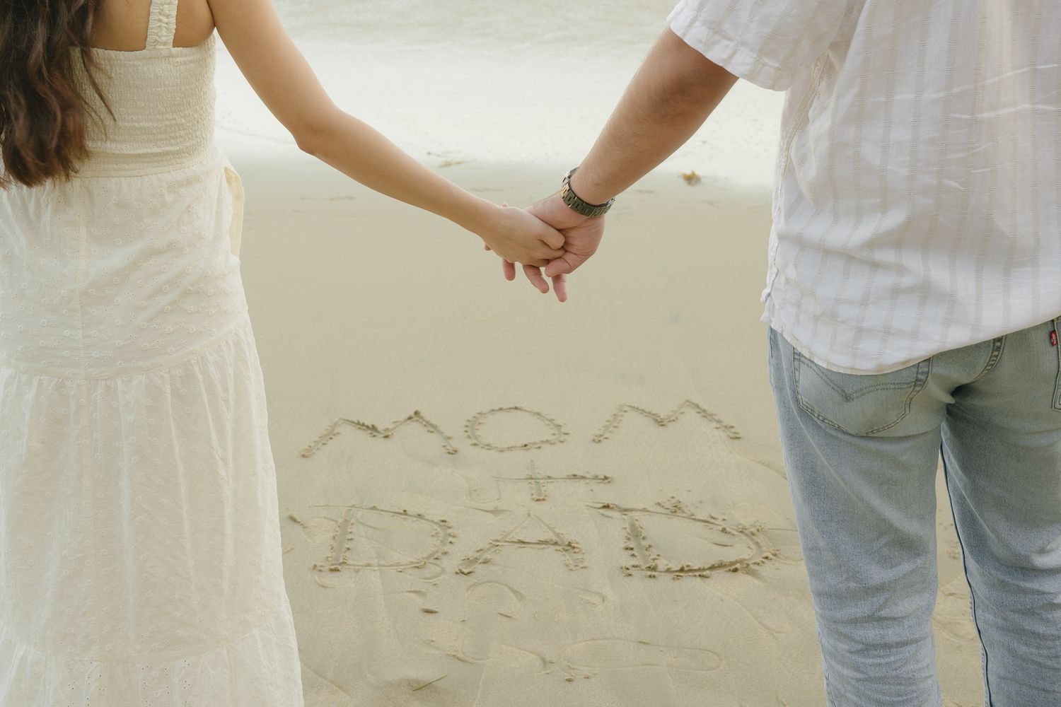 Couple holding hands on beach with Mom and Dad written in sand, romantic outdoor photo.