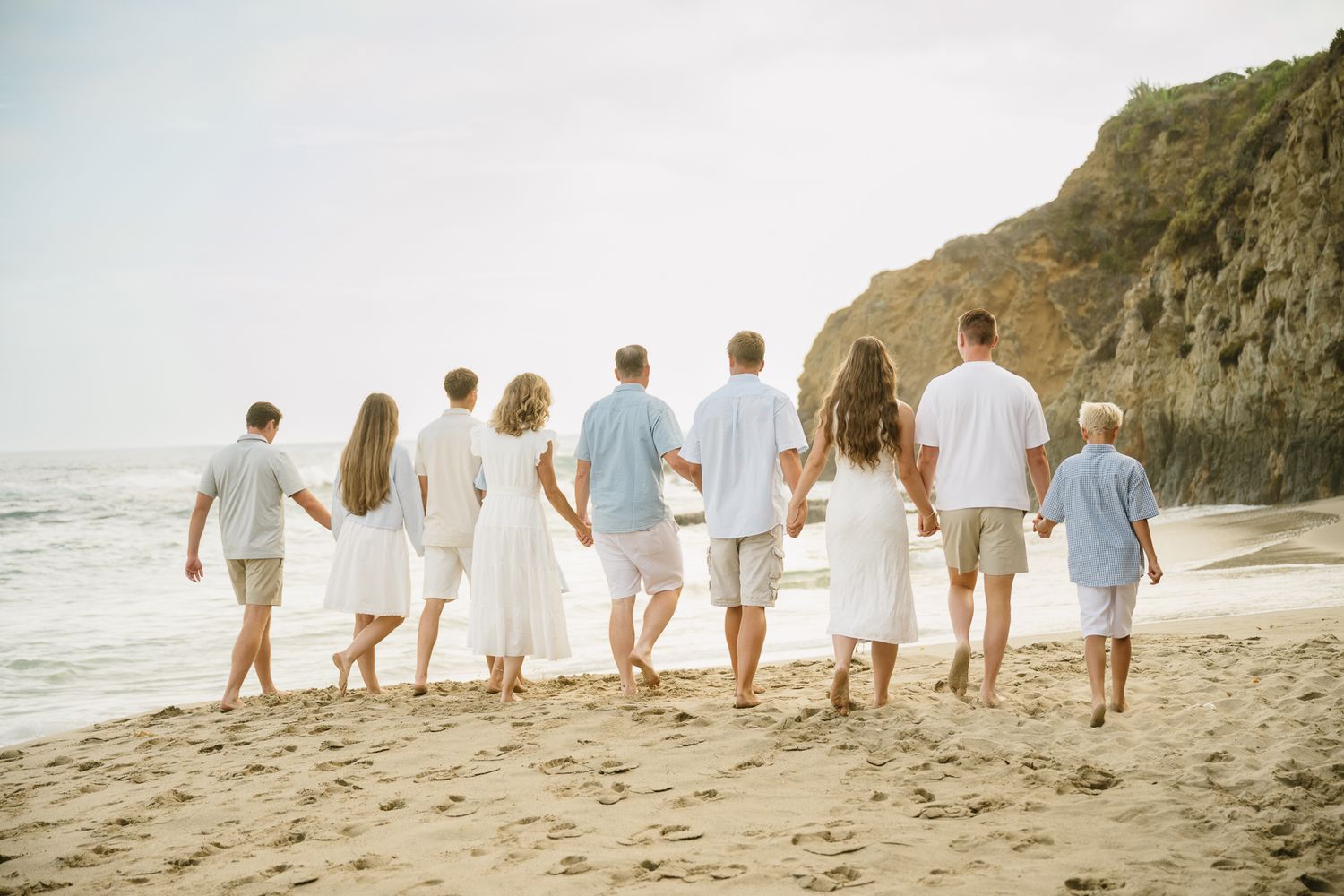 Family of eight walking hand-in-hand on a sandy beach in white and blue outfits with ocean waves and cliffs behind.