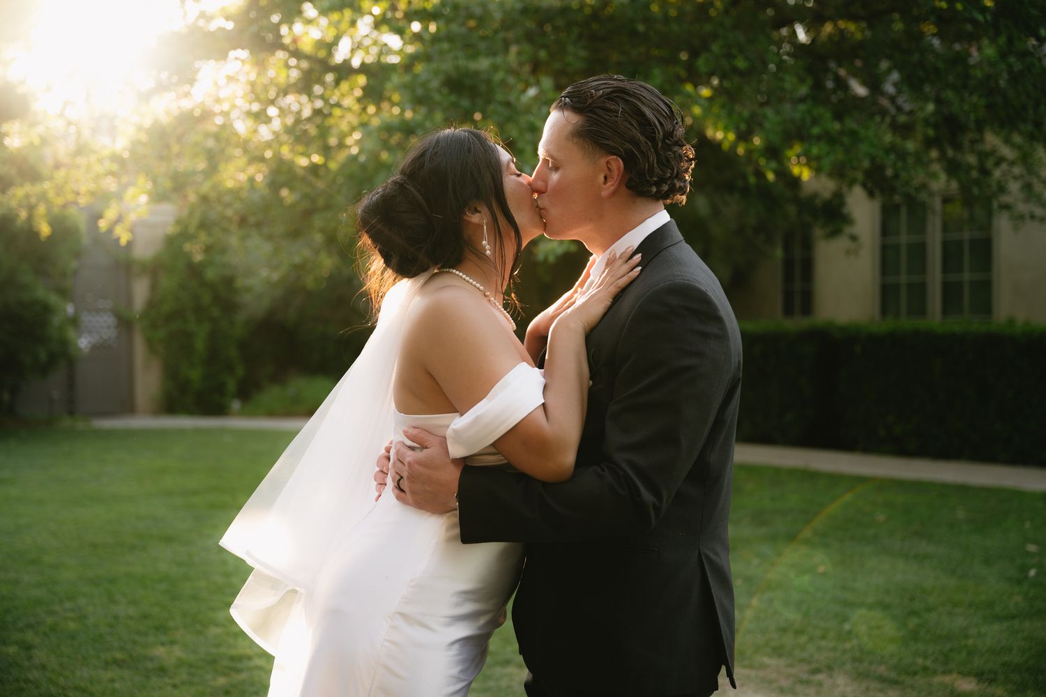Bride and groom share a romantic kiss outdoors at golden hour, surrounded by lush greenery.