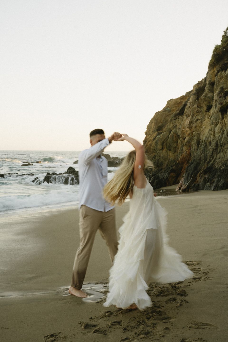 A couple dances barefoot on a sandy beach, the bride in a flowing white gown, with rocky cliffs and ocean waves behind them.