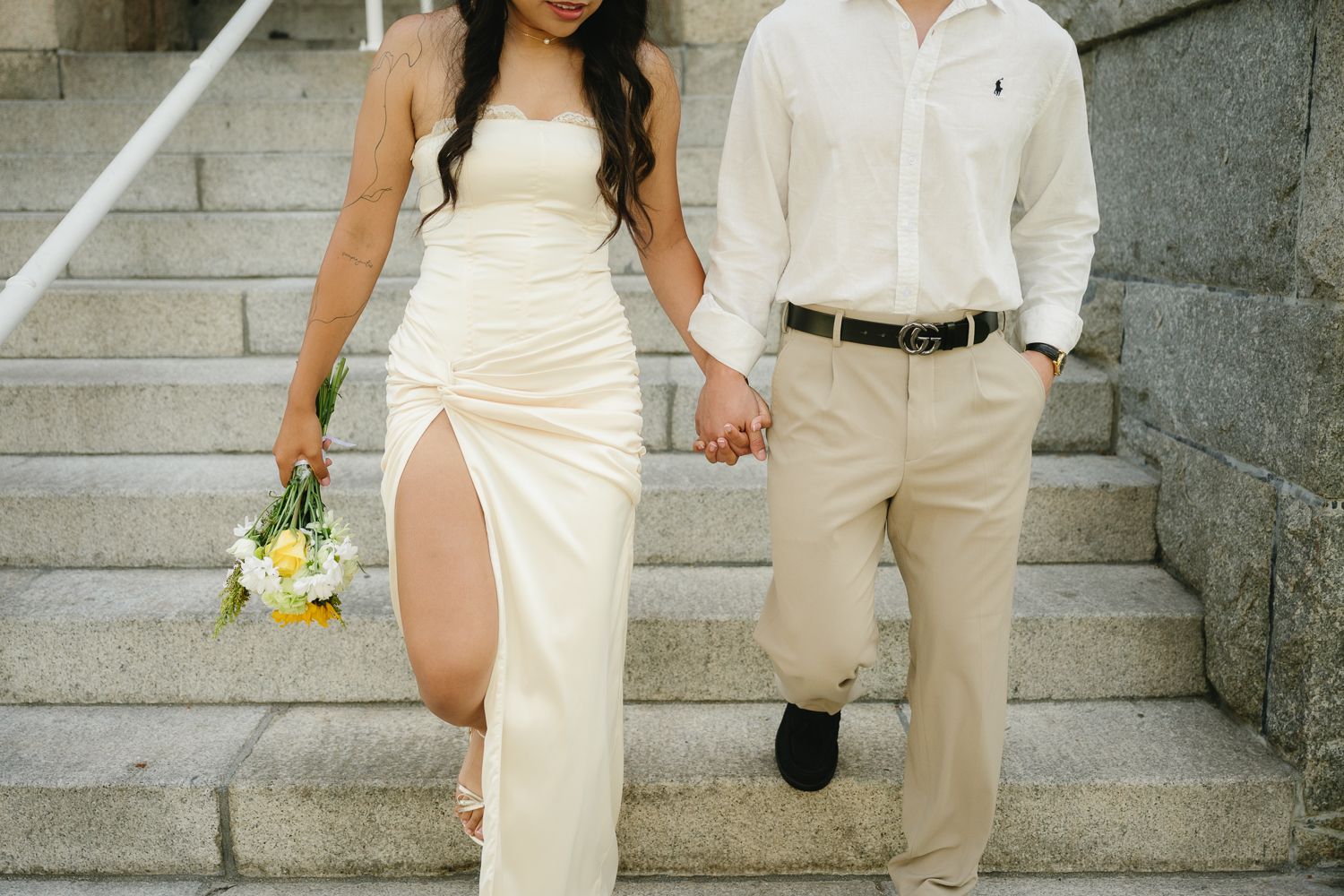 Couple holding hands on stone steps, woman in white slit gown with bouquet, man in white shirt and khaki pants.