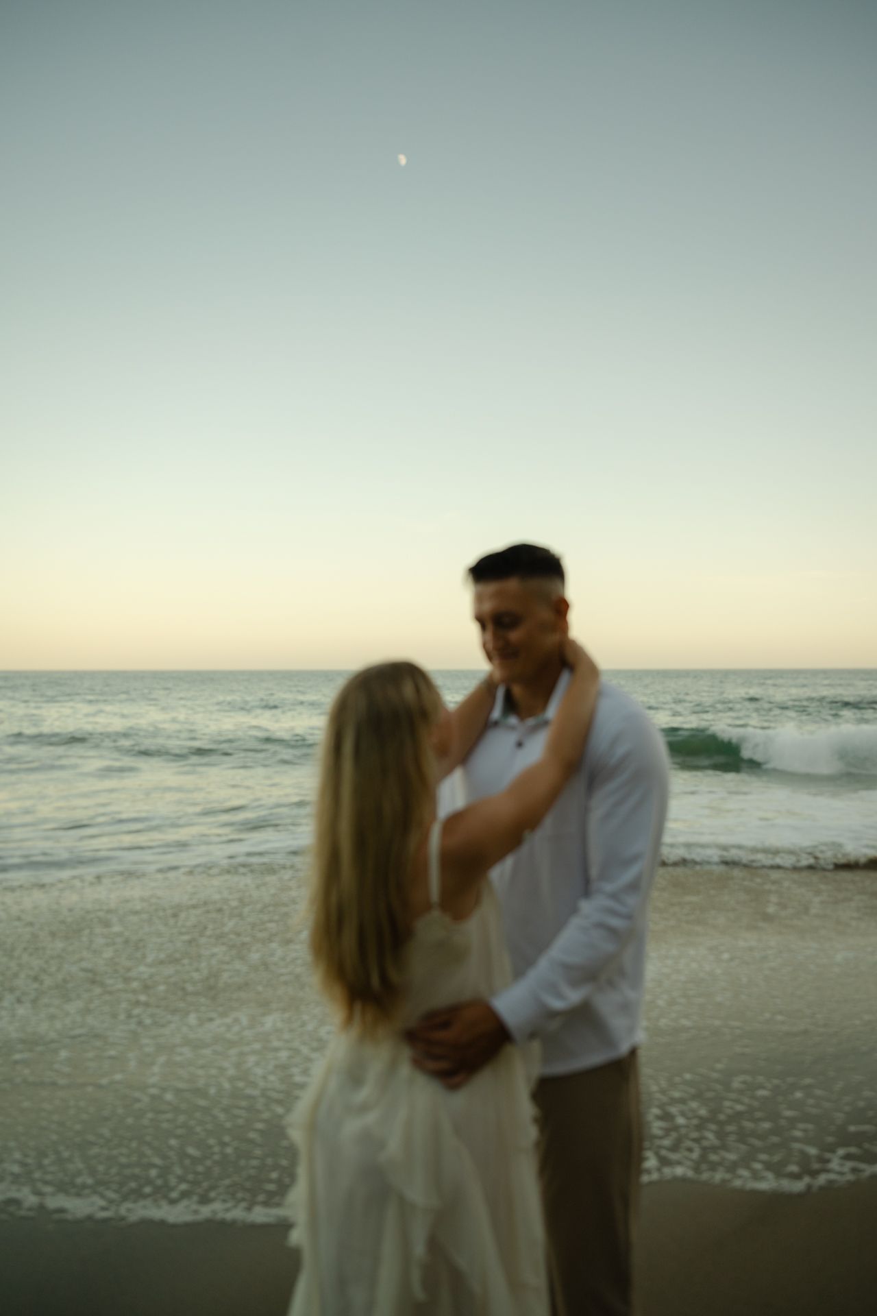 Couple embracing on beach at sunset, woman in white dress touching mans face, ocean waves and crescent moon in background.