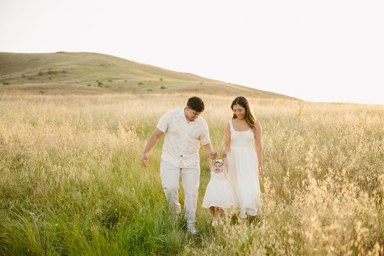 Family in white outfits walking through golden meadow grass with rolling green hills in warm sunset light.