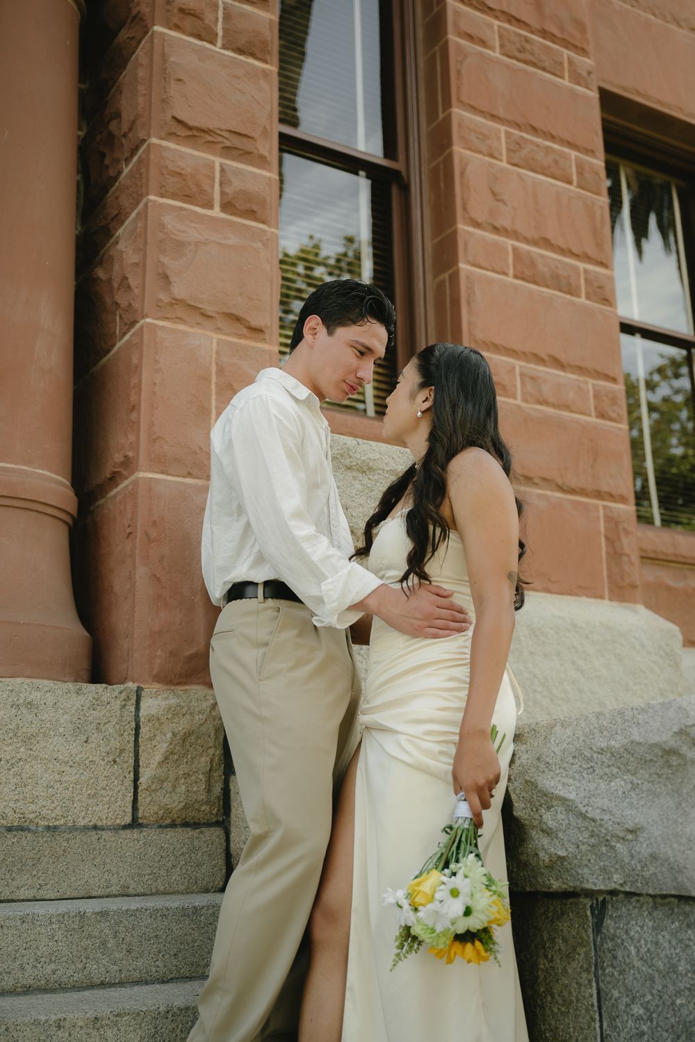 Bride and groom share a romantic moment on stone steps outside a historic brick building, bride holding yellow and white bouquet.