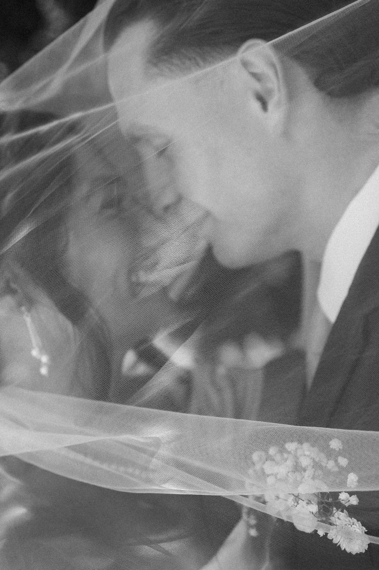 Black and white photo of a bride and groom sharing a tender moment under a sheer wedding veil with delicate flowers.