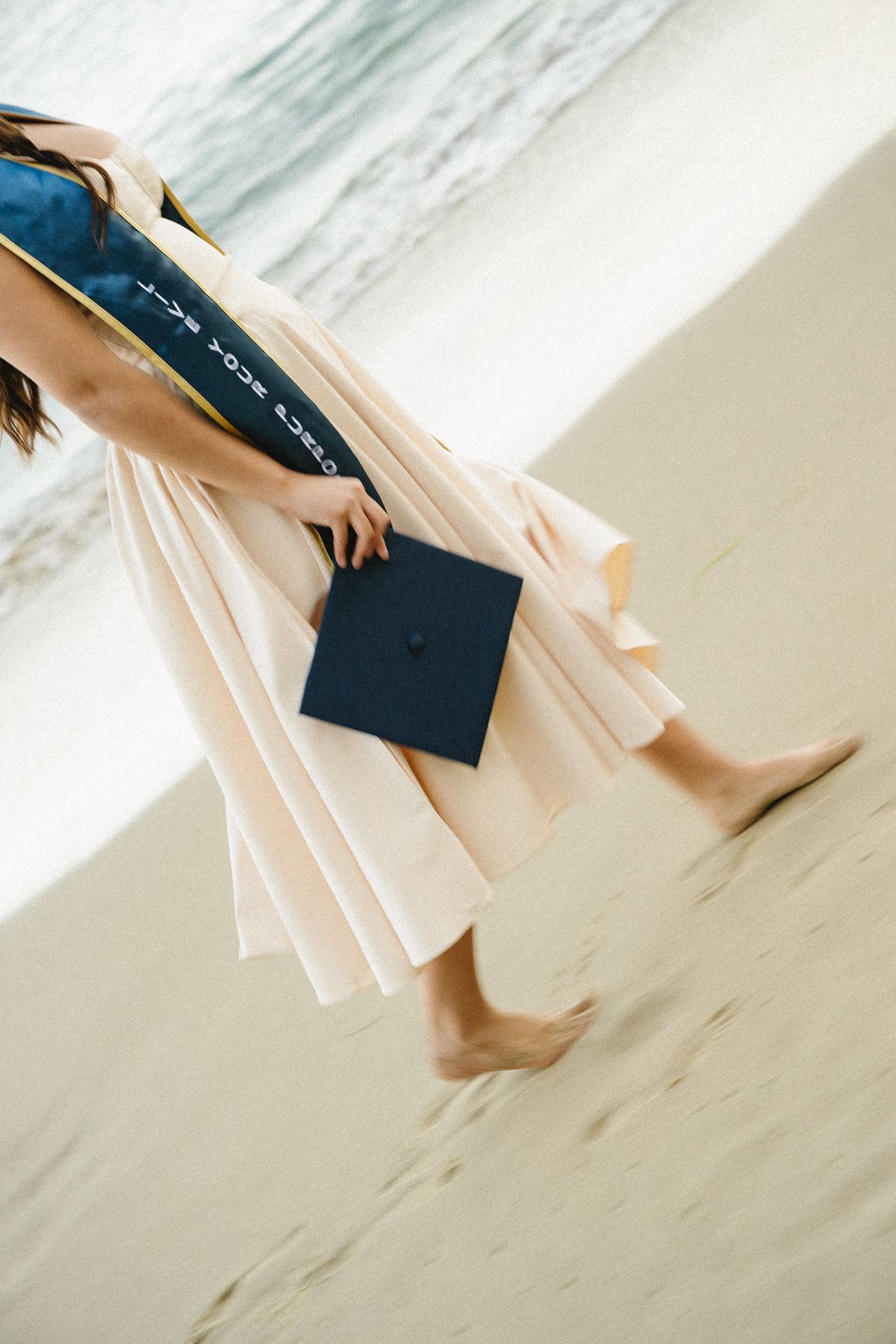 Graduate in white gown holding mortarboard cap walks barefoot on sandy beach near ocean waves.