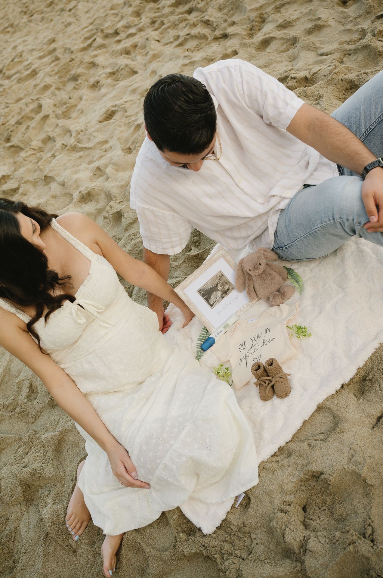 Couple sitting on sandy beach with baby items and ultrasound photo, maternity announcement session.