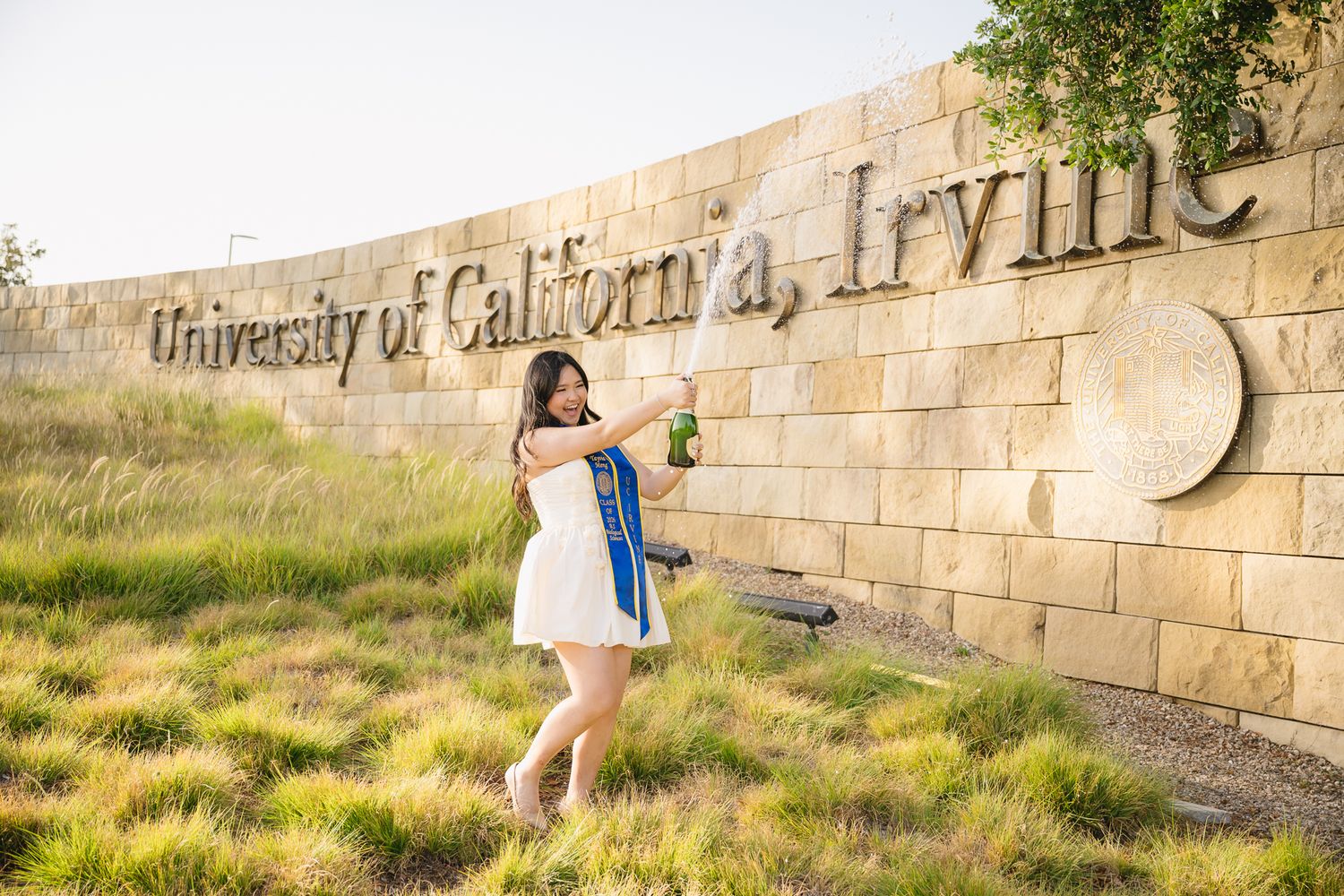 Graduate poses in white dress with blue sash in front of University of California, Irvine stone sign.